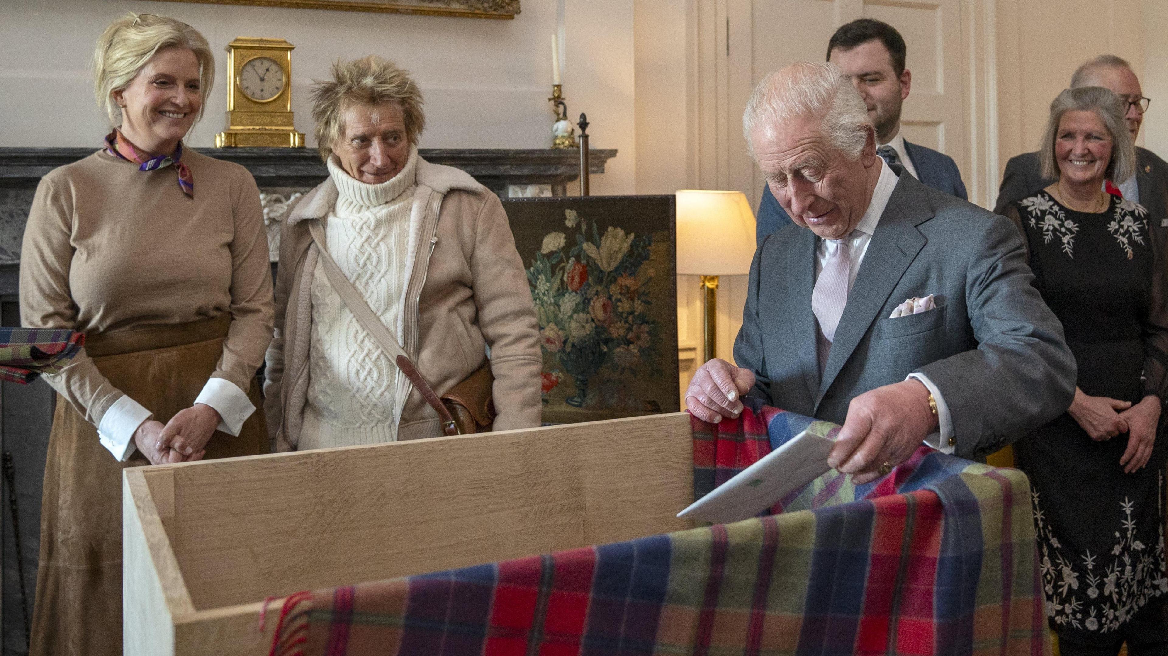 King Charles, wearing a grey suit with a white shirt and light tie, on the right of the image placing a white letter into a large wooden box with a tartan blanket partially covering it. It is taking place indoors. Sir Rod Stewart, wearing a white turtleneck jumper under a beige jacket and with a beige strap covering his torso, and wife Penny Lancaster, wearing a beige dress, watch on.