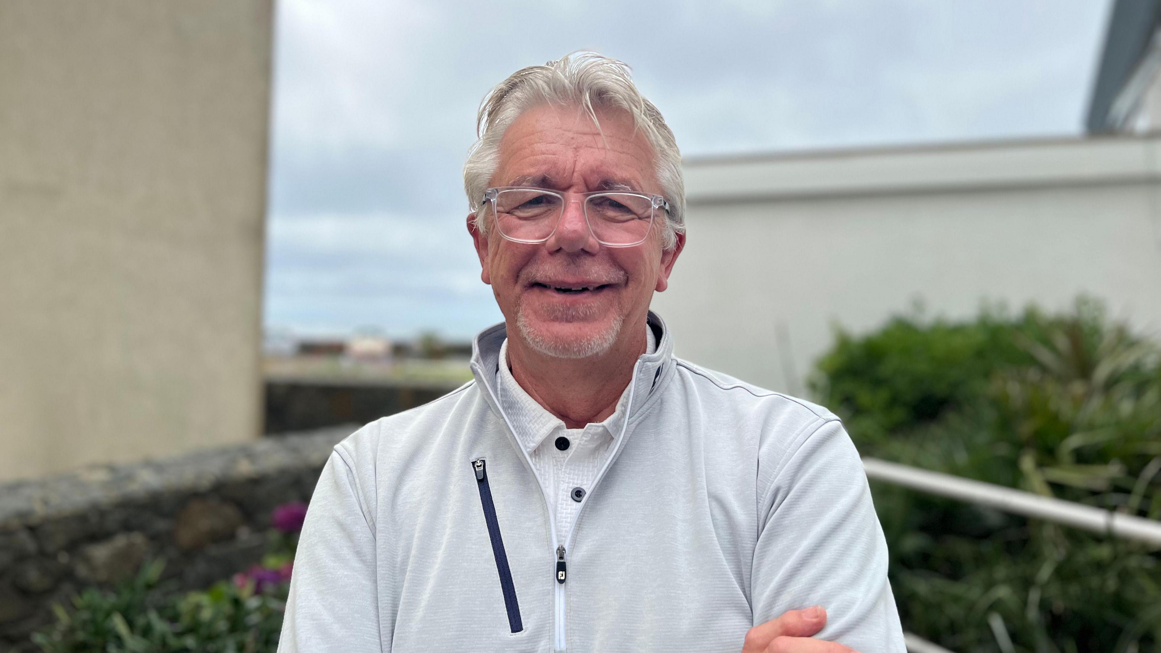 A headshot of a man with grey hair smiling at the camera. He's wearing clear glasses a white collared shirt with a grey quarter zip over the top.