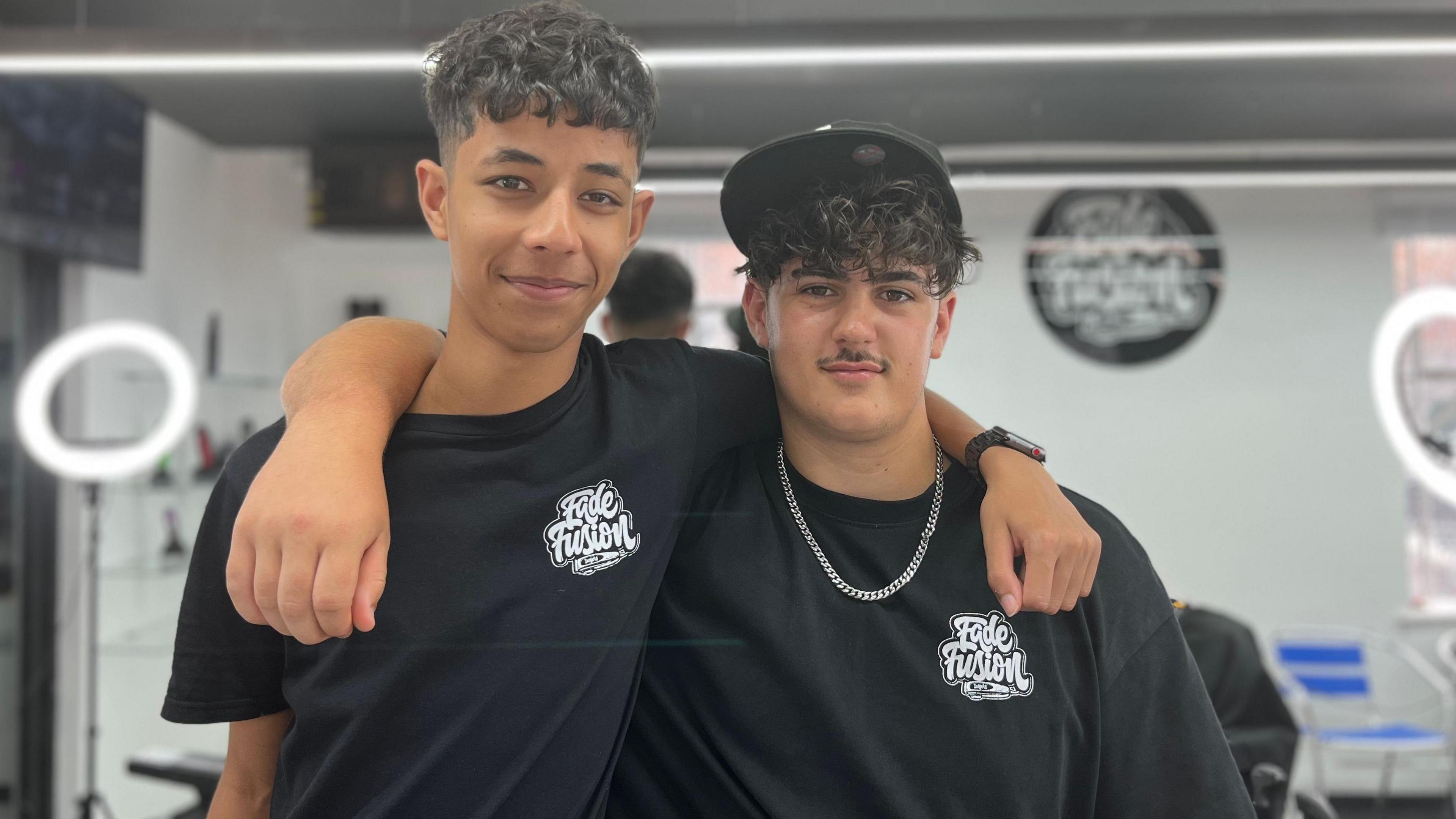 Two boys are wearing black t-shirts and have their arms over each others shoulders. They are standing in the barber shop with white ring lights in the background.