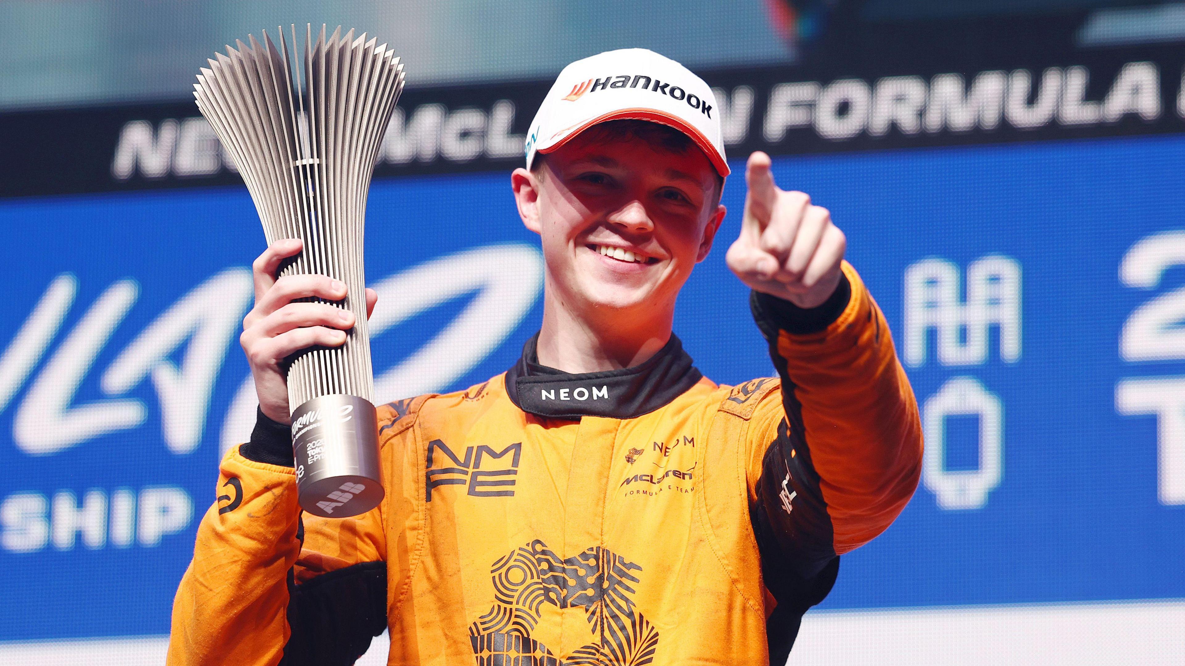 Taylor Barnard wears an orange racing jumpsuit. His hand is outstretched and finger pointing out. In his other hand he is holding a silver trophy. 