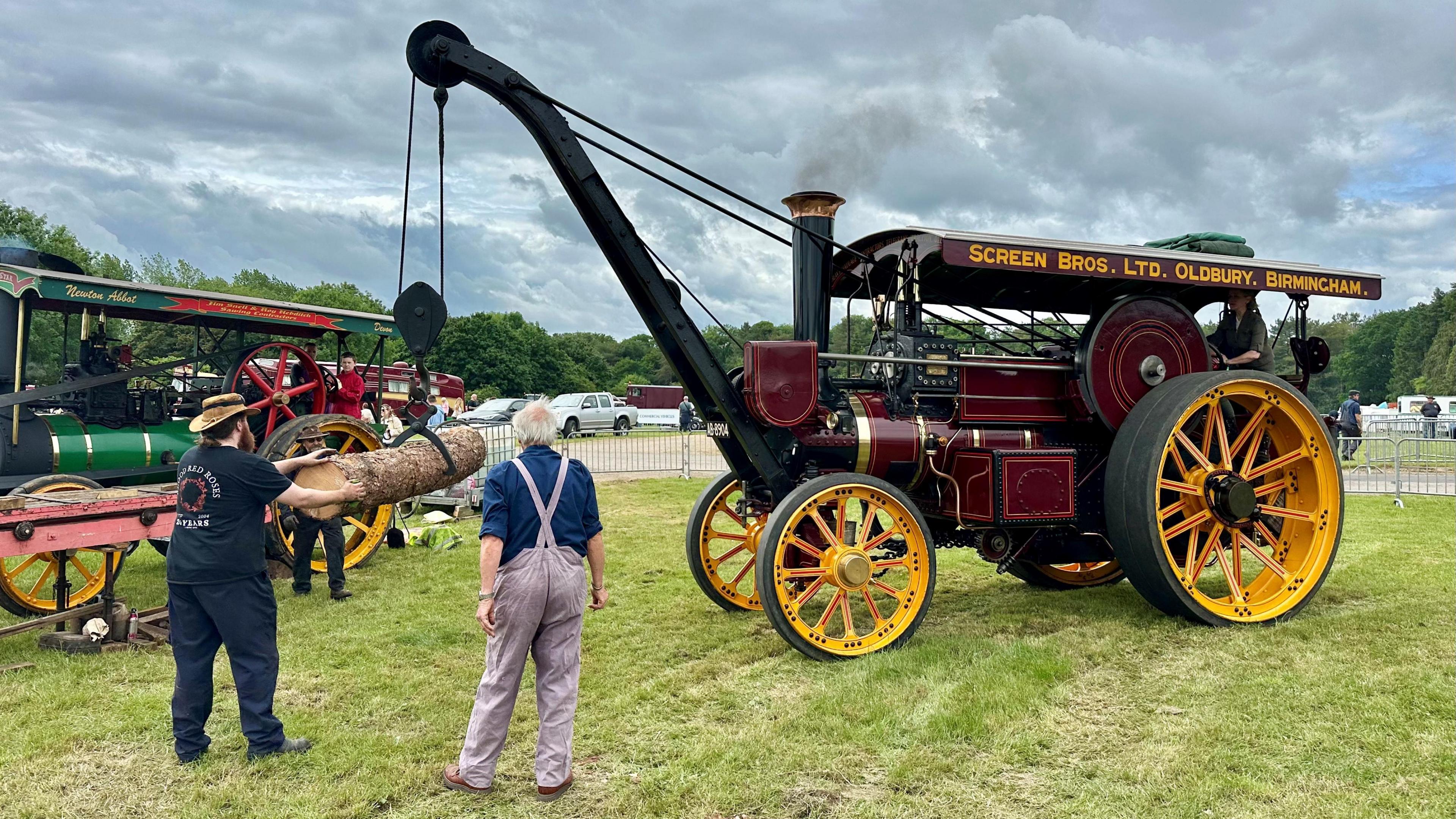 A steam engine with a rudimentary crane lifts a log, which is being guided by a man wearing a hat with a further steam engine and a couple of other people in the background.