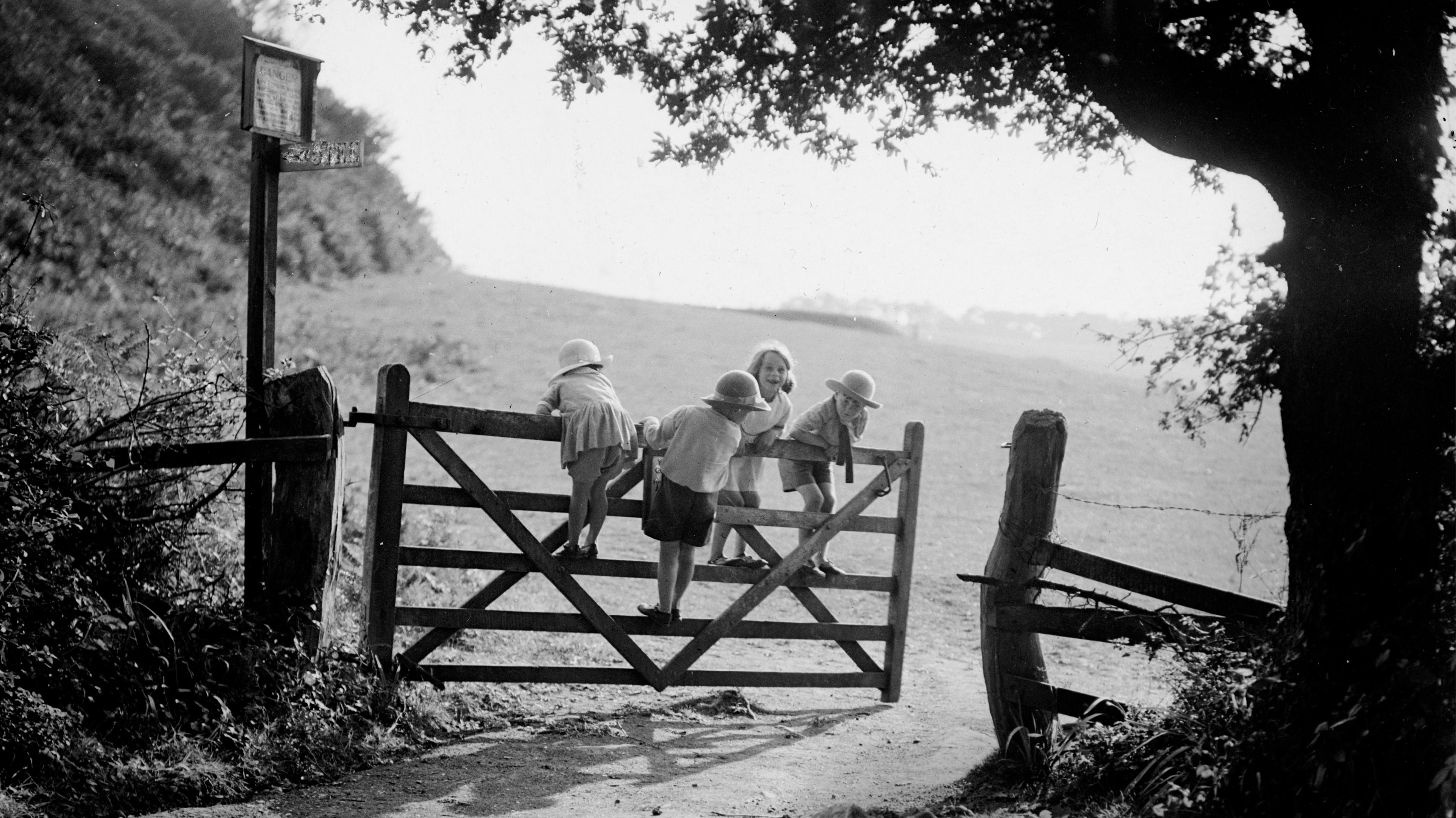 A black and white photo of four young children standing on a gate to a field. It looks like a sunny day as they are wearing skirts and shorts and hats.