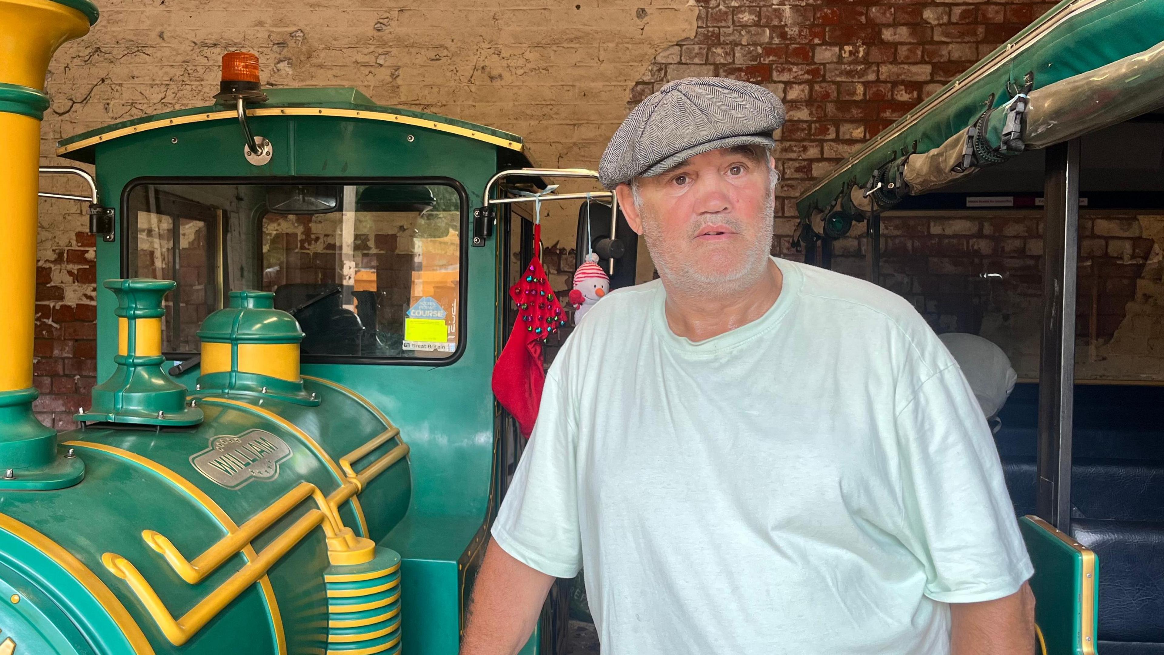 A man wearing a tweed cap and a white T-shirt stands inside a brick building. He is standing next to a green and yellow land train on his right and a green carriage on his left.