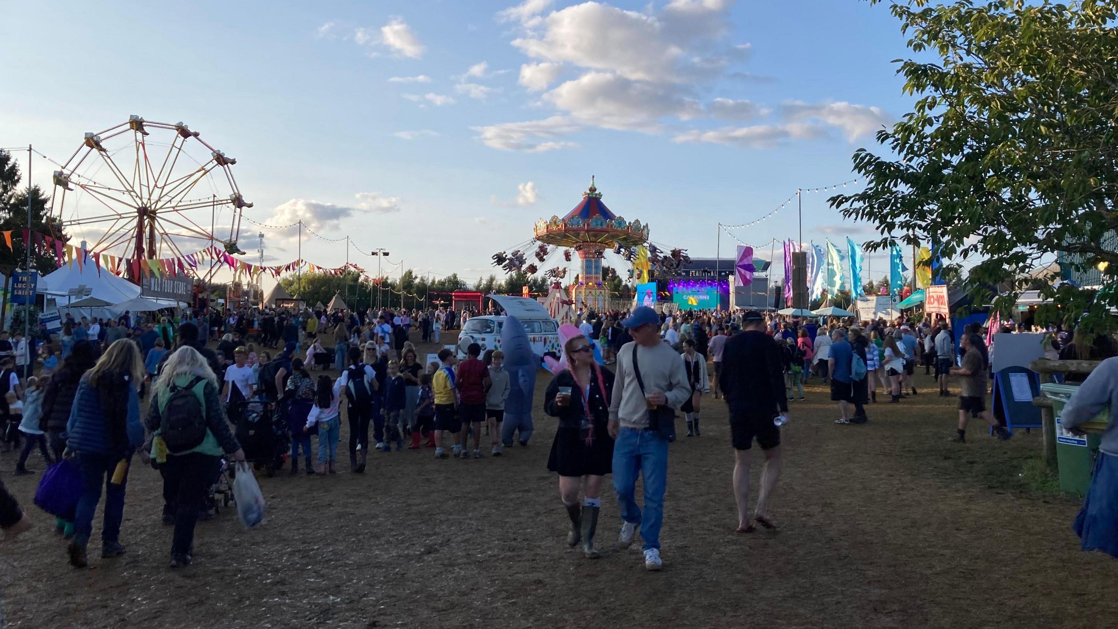 The Big Feastival pictured in 2024. It shows people walking on muddy grass with fairground rides and flags in the background.