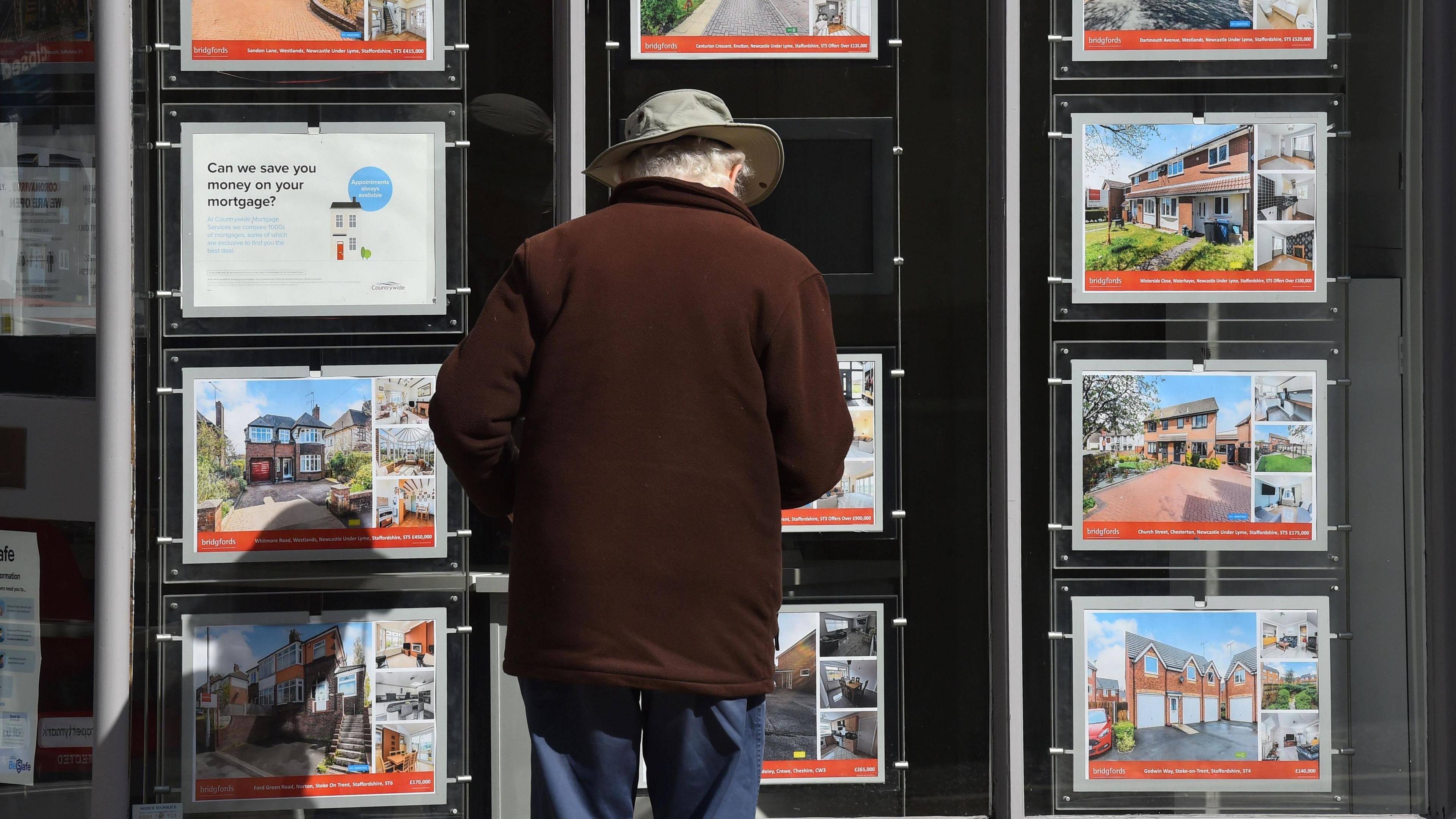 An elderly man looks at signs for renting and buying houses outside an estate agent's shop. He's wearing a hat and a brown coat.