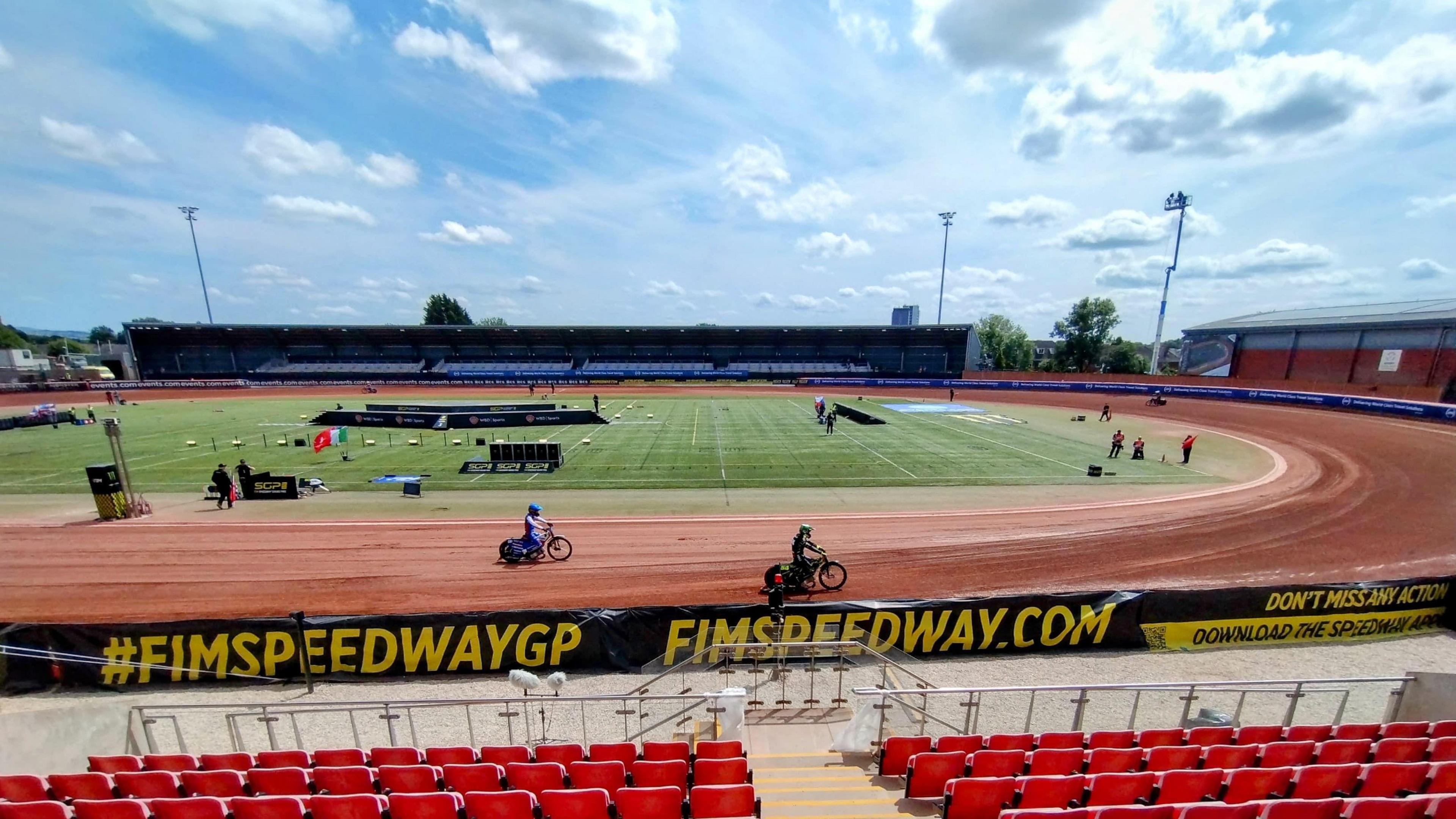 A race track with red seating in front. Motorcycles can be seen on the red, dirt, track. There is a circular grass patch in the middle of the track. It is a sunny day with blue skies. 