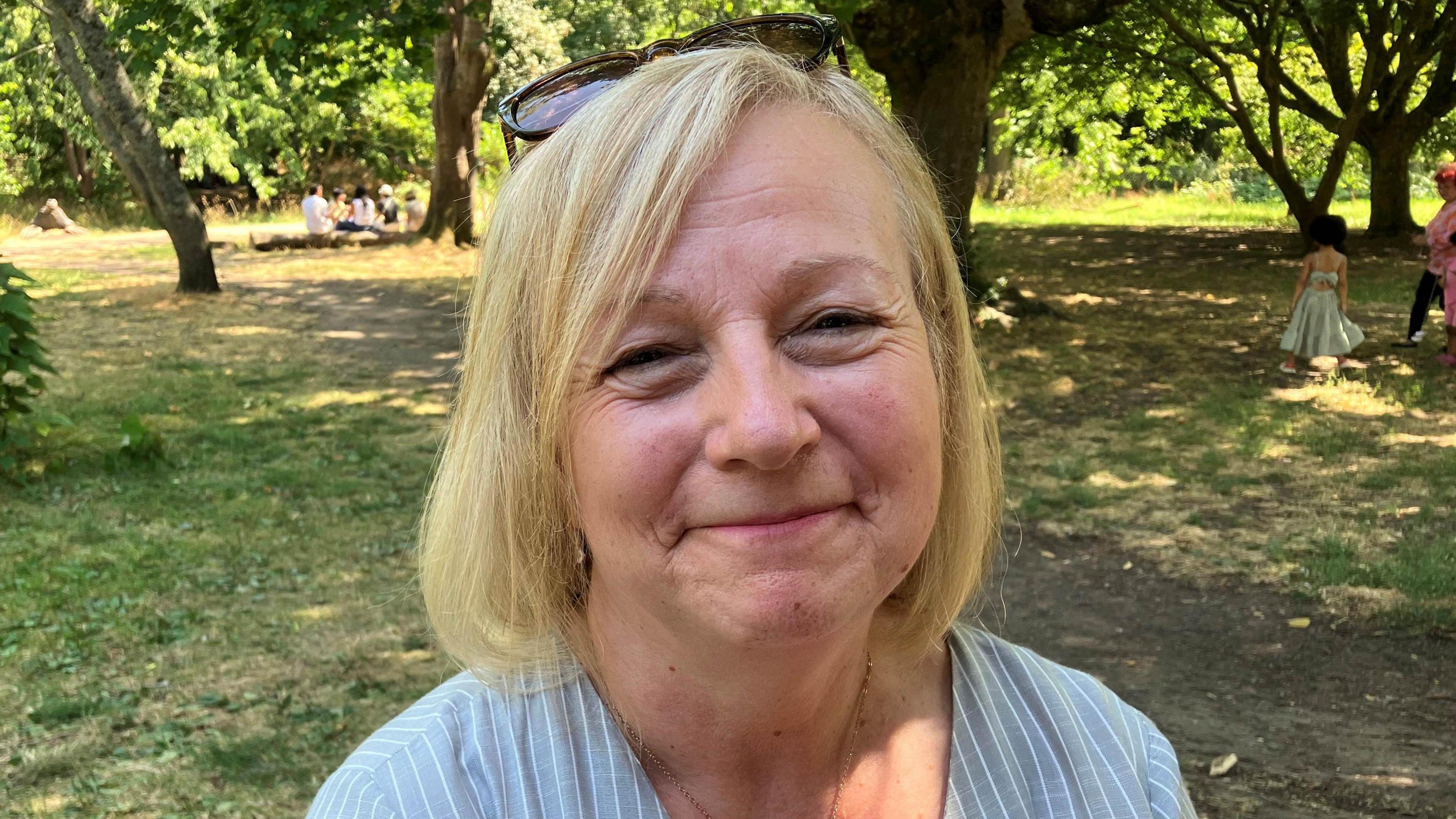 Melani Smith is photographed outside in a park. Grass and trees can be seen behind her. She has short shoulder length blonde hair and is wearing sunglasses on top of her head. She is smiling and looking at the camera.