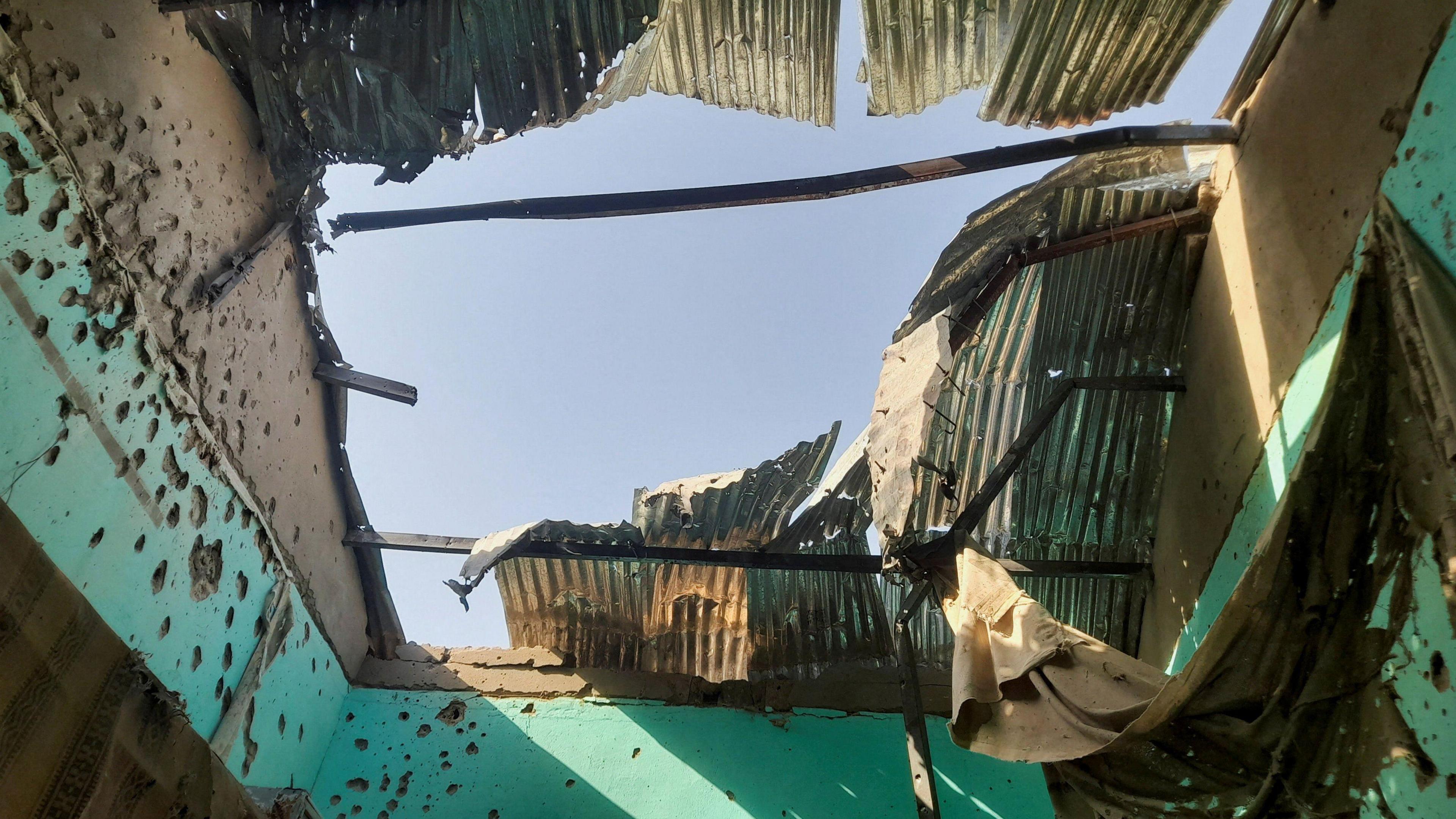 A gaping hole in the roof of a building, with the remains of corrugated iron sheets and bullet marks in the walls.