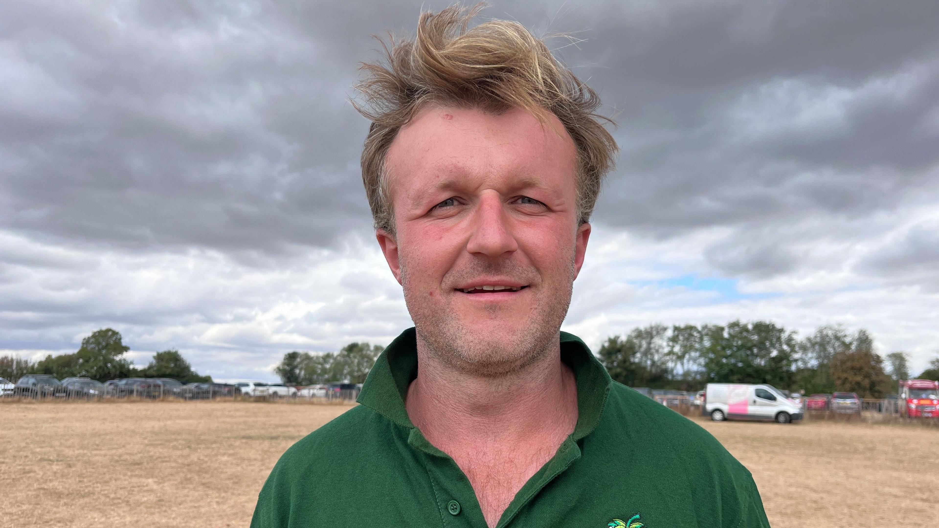 A man in a green polo shirt is stood in a field with grey clouds above. He is smiling and looking directly at the camera. He has blond hair. 