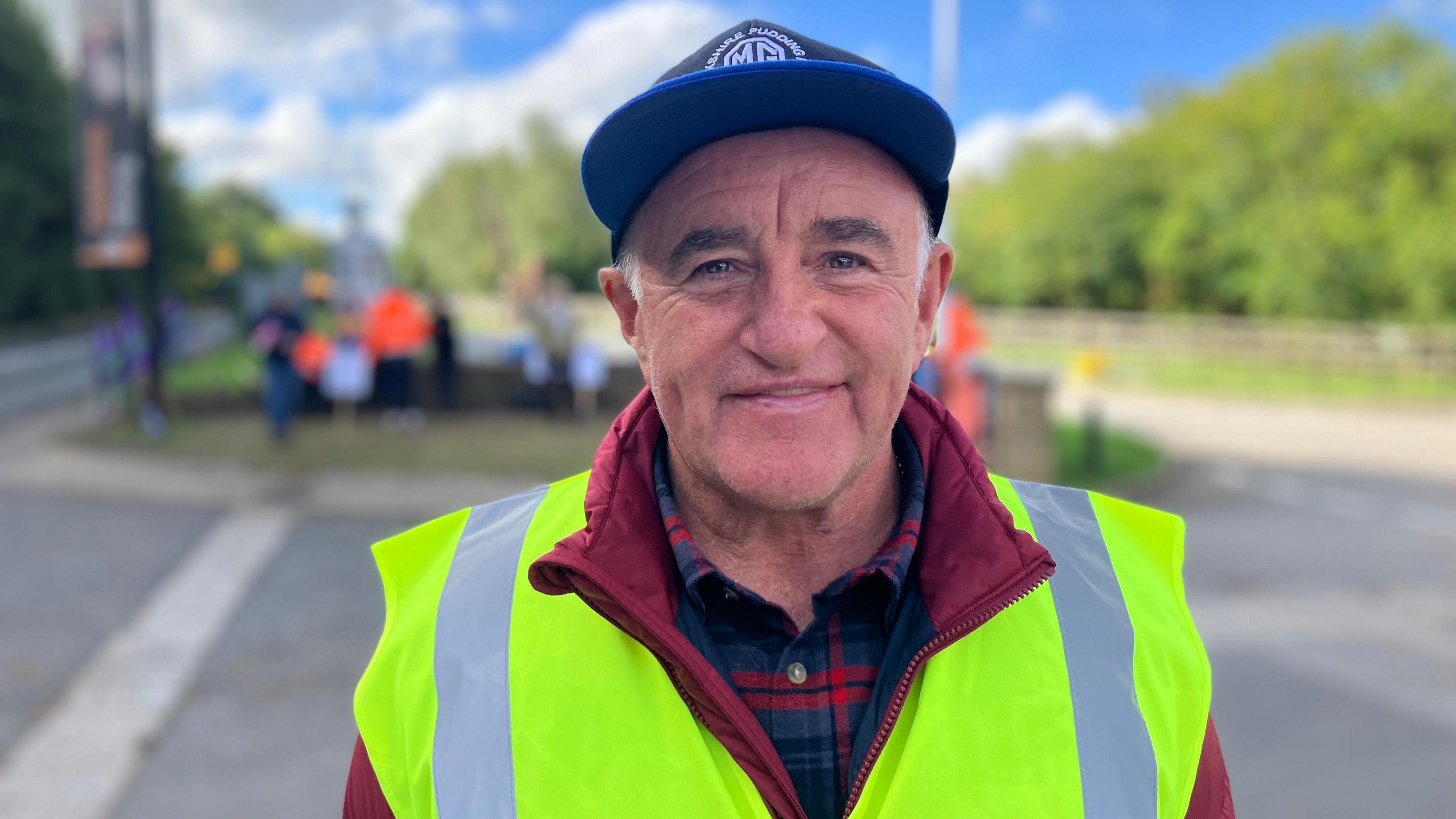 A man wearing a blue cap and a yellow high vis jacket smiles at the camera. Behind him, a crowd of picketers are blurred.