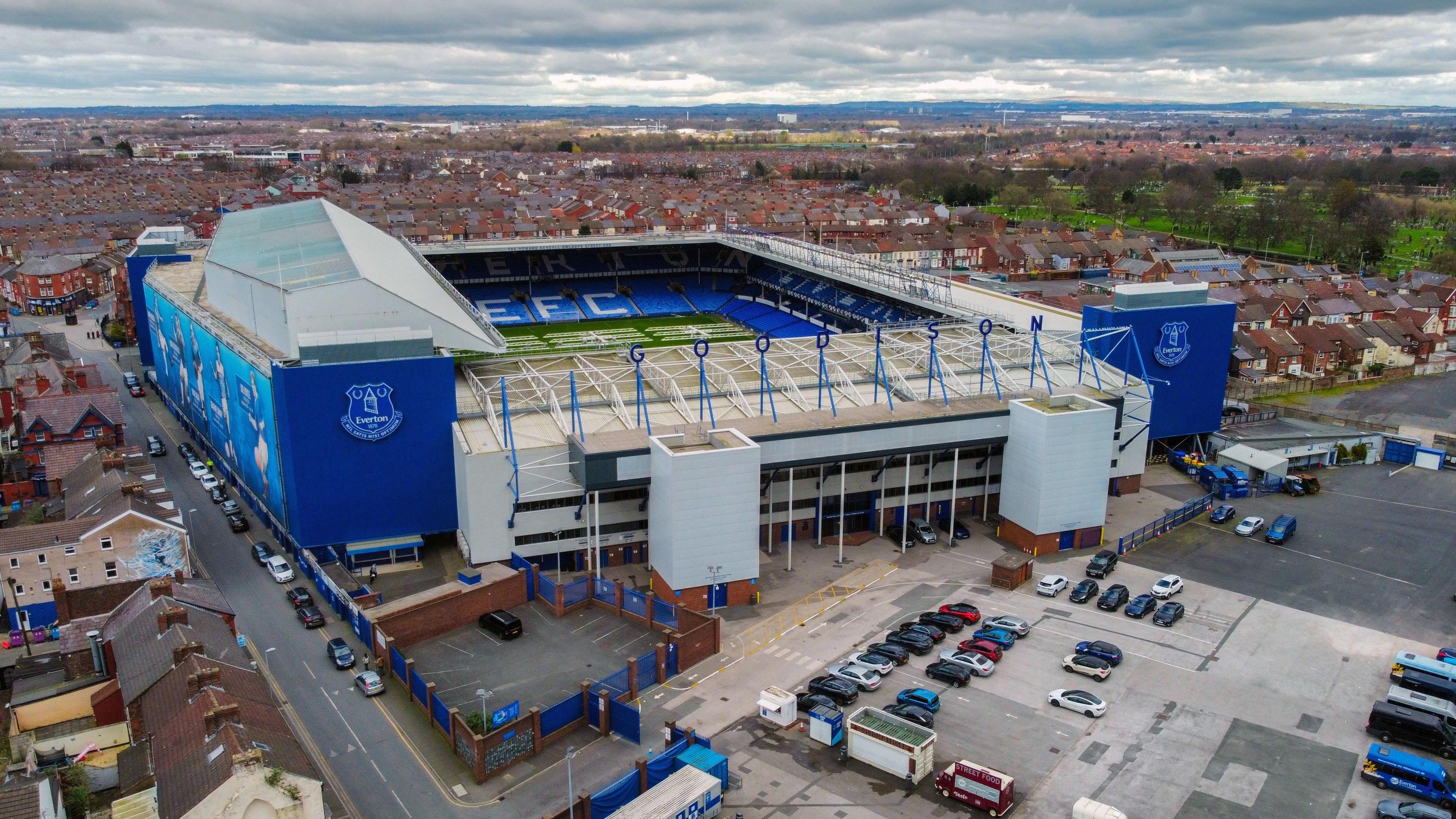 An aerial view image captures Goodison Park from up high. There is a car park situated at the front with housing surrounding the rest of stadium which is royal blue and white.