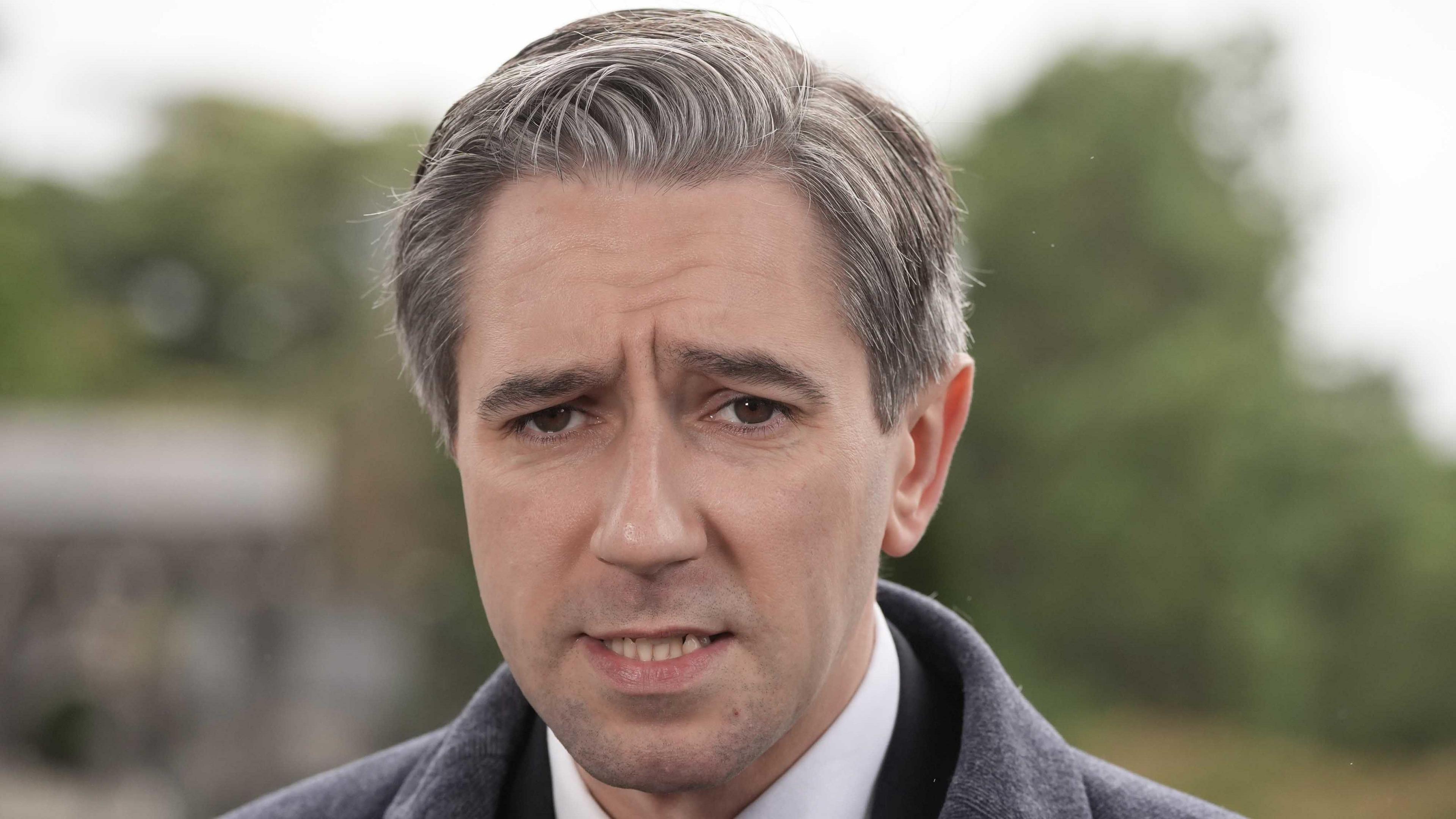 Simon Harris - a man with grey hair stands in front of a blurred background. He is wearing a black suit jacket, a white collared shirt and a red and white polka dot tie.