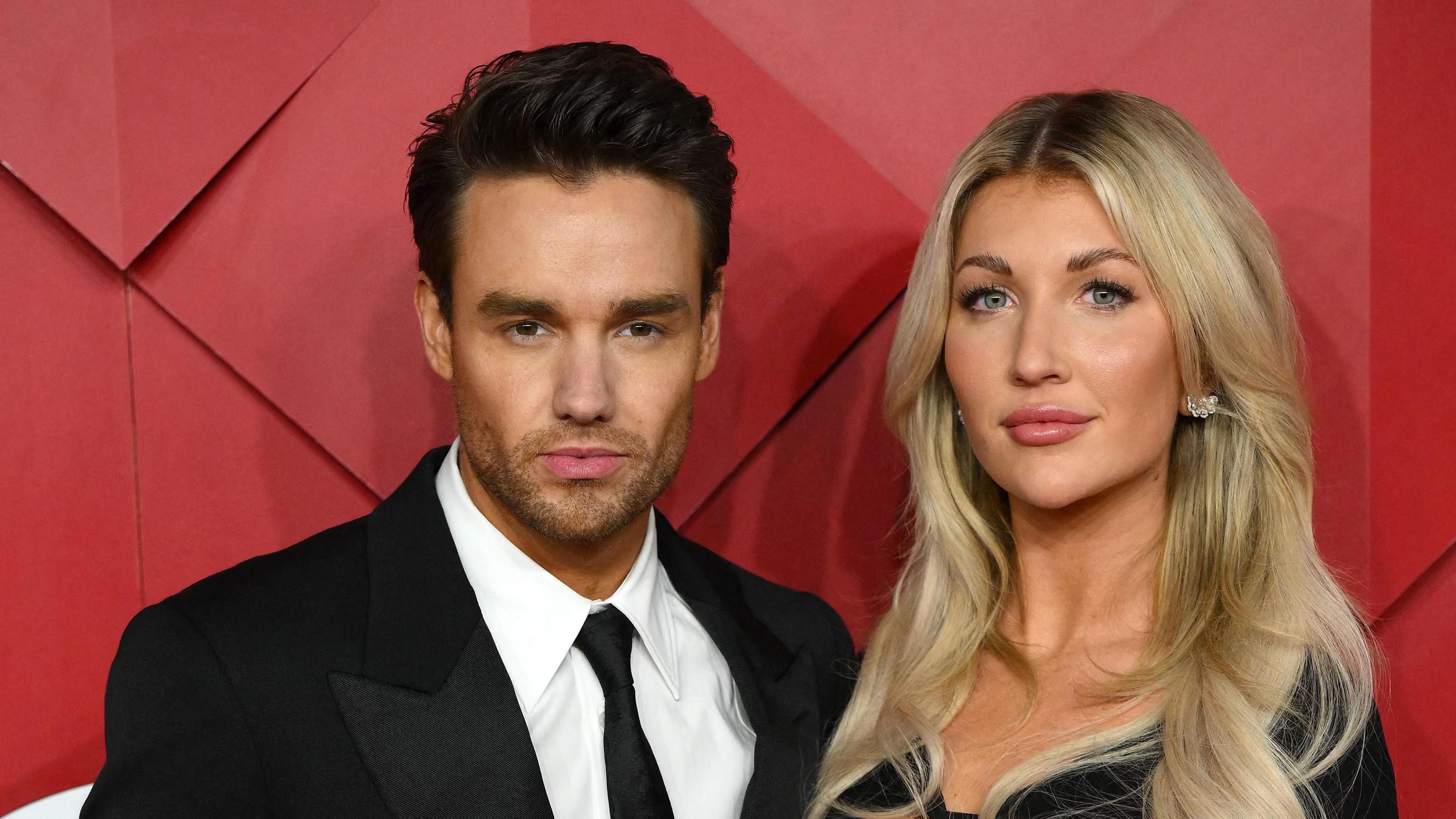 Liam Payne in suit and tie stands beside a smiling Kate Cassidy, with blonde hair and glittery earrings, against a red background