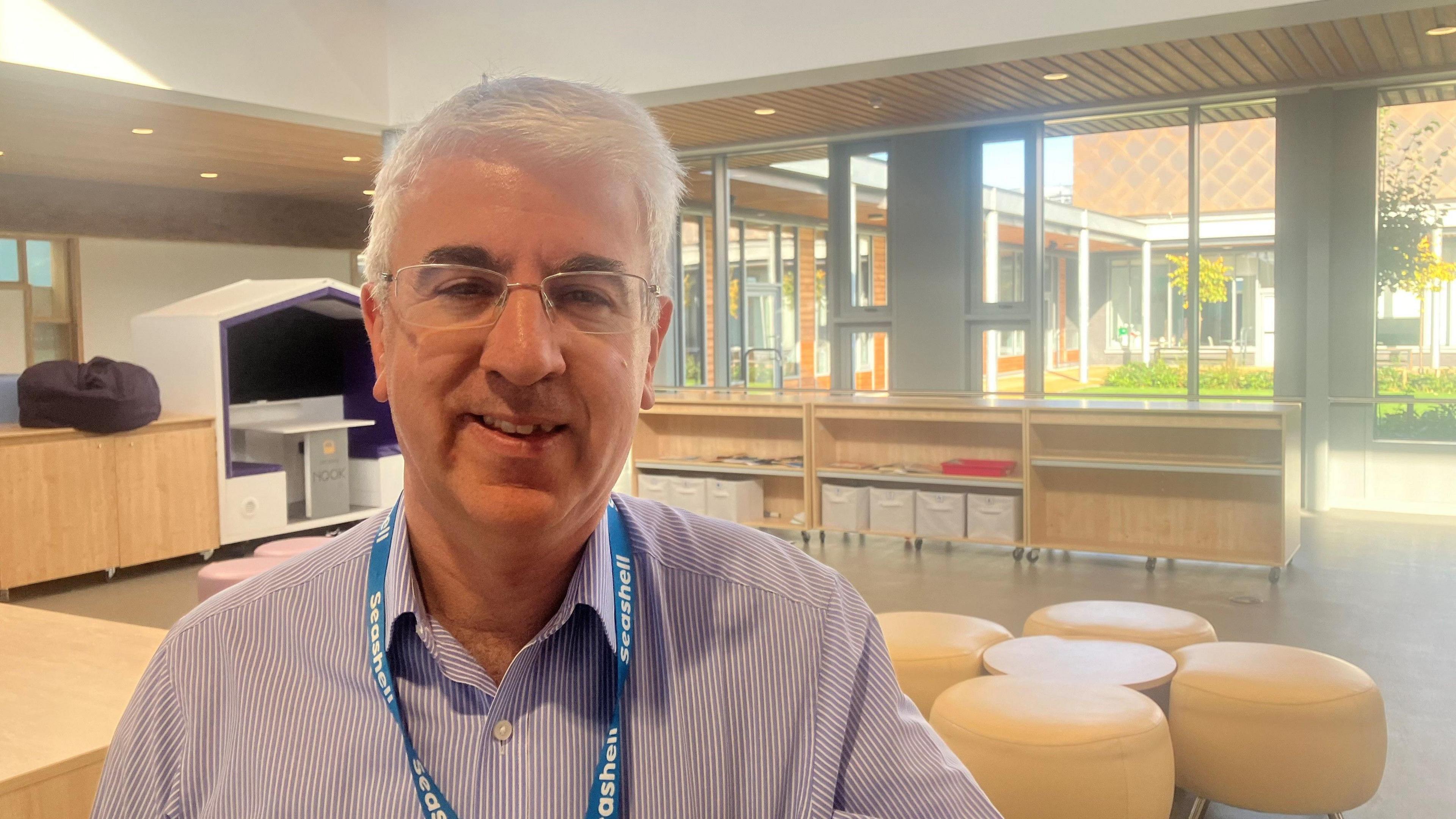 Photo of Brandon Leigh of the Seashell Trust inside the organisation's new building. He is looking directly at the camera and behind him is a seating area containing beige coloured stools, and various cupboard units. 