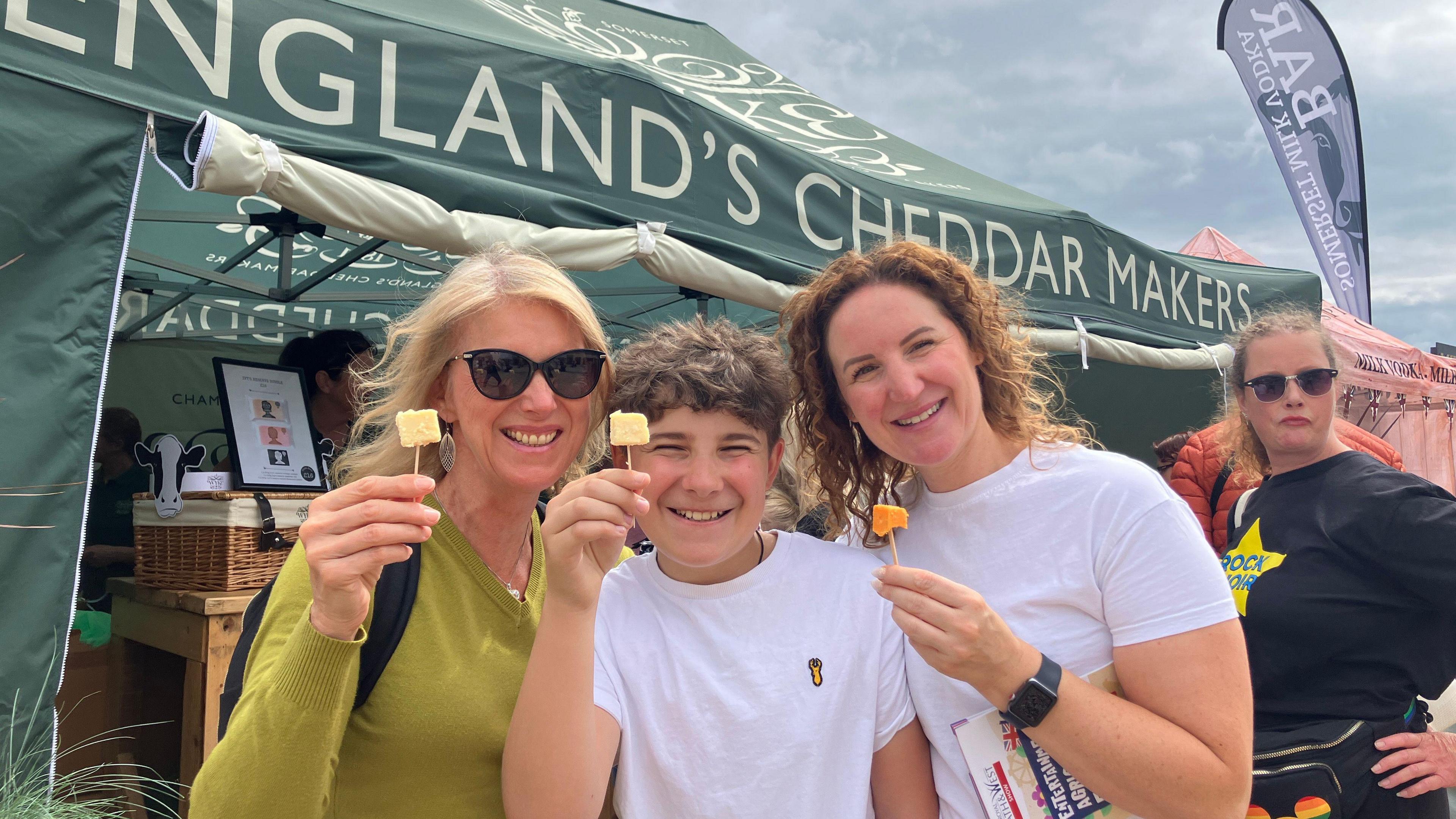 Two women and a boy stand gathered together looking at the camera smiling as they hold small pieces of cheese on sticks at the Bath and West Show.