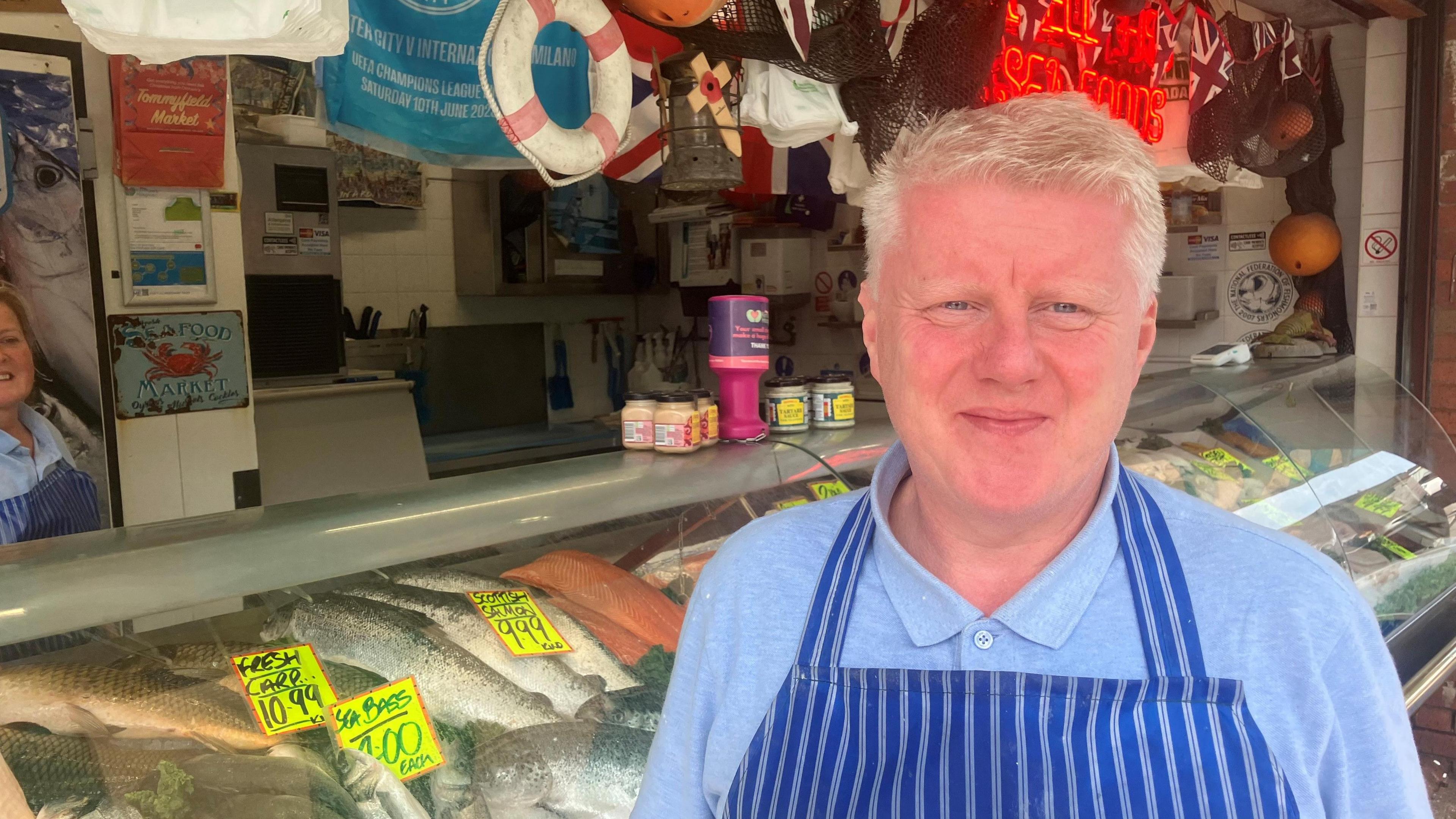 Photograph of Mark Crossley in front of his fishmonger's stall at Oldham's Tommyfield Market. In the background are his fish.
