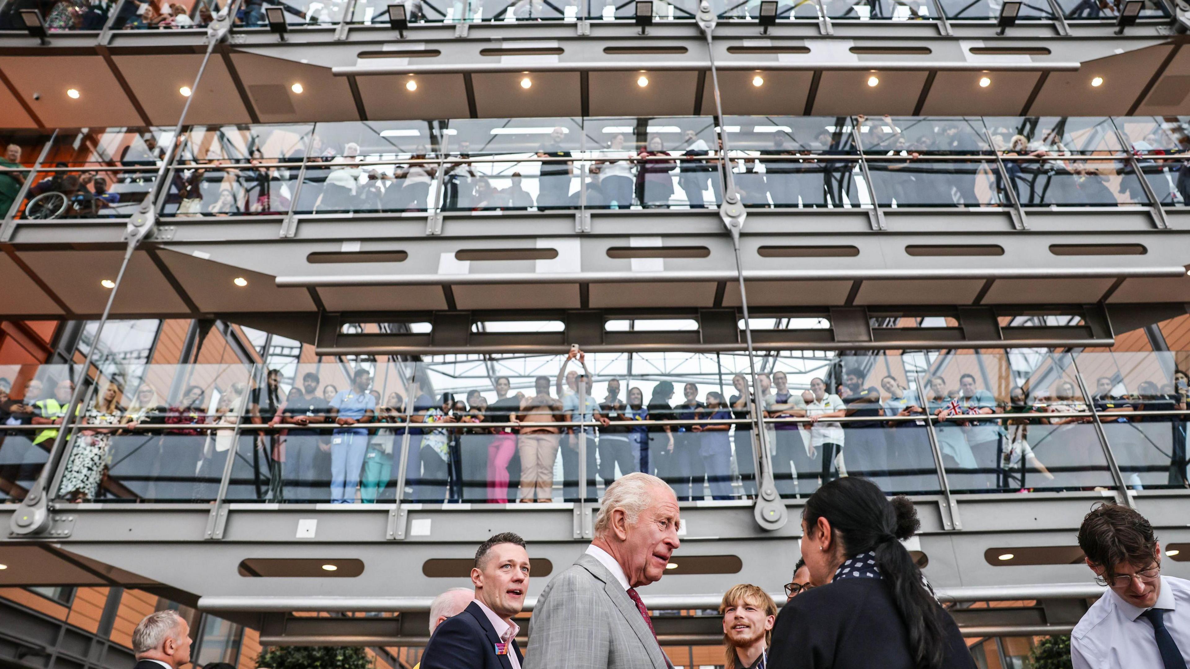 Three levels of glass walkways viewed from ground level inside an internal atrium, each lit by spotlights, with crowds looking down onto the King on the ground level as he chats to groups of people.