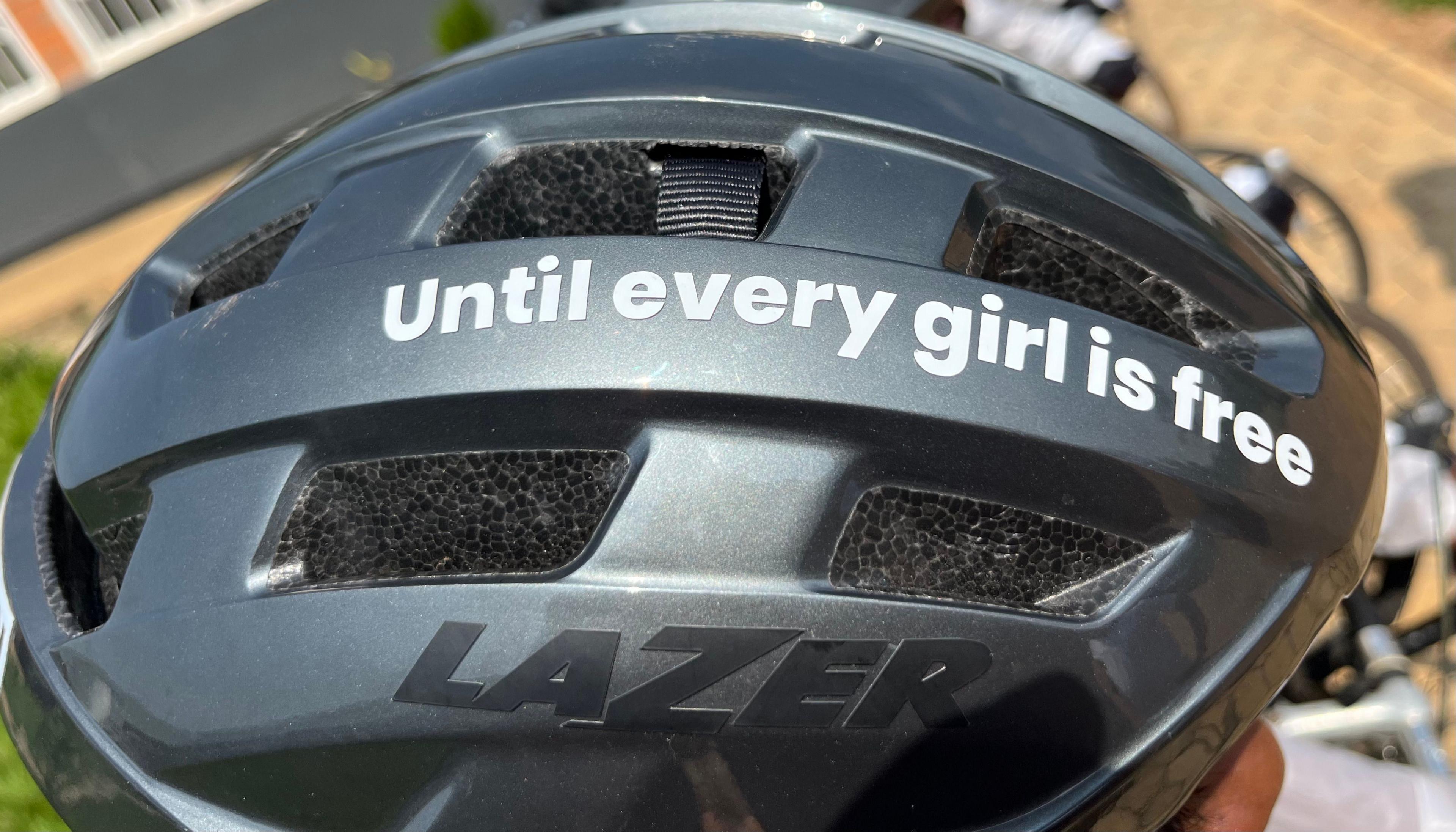 Detail of a grey cycling helmet seen from above bearing the message 'Until every girl is free' in white text running down its middle from back to front