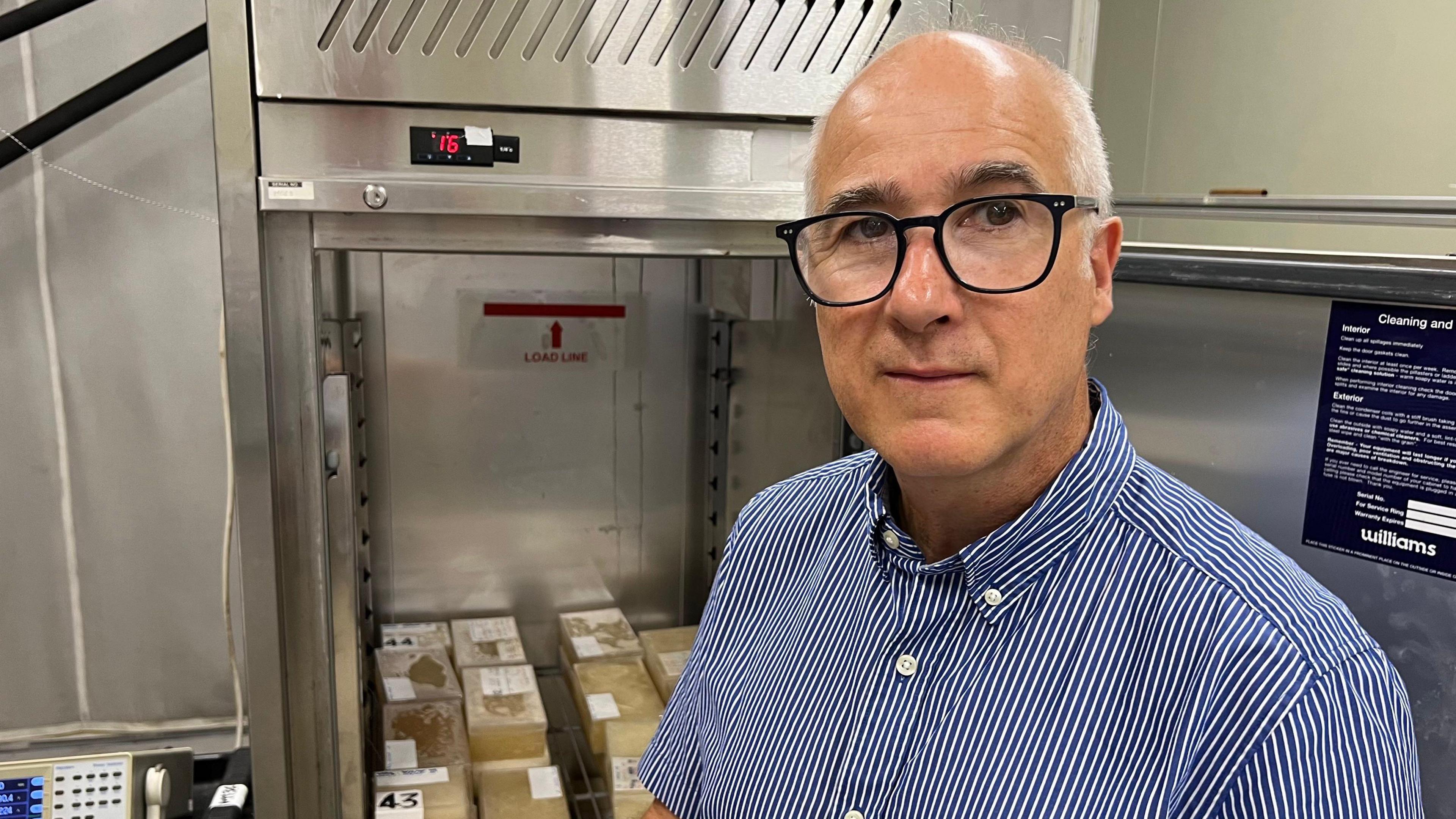 A man in a blue striped shirt stands in front of a stainless steel fridge. The door is open, showing it is full of blocks of cream coloured gel. There are wires coming out of the blocks.