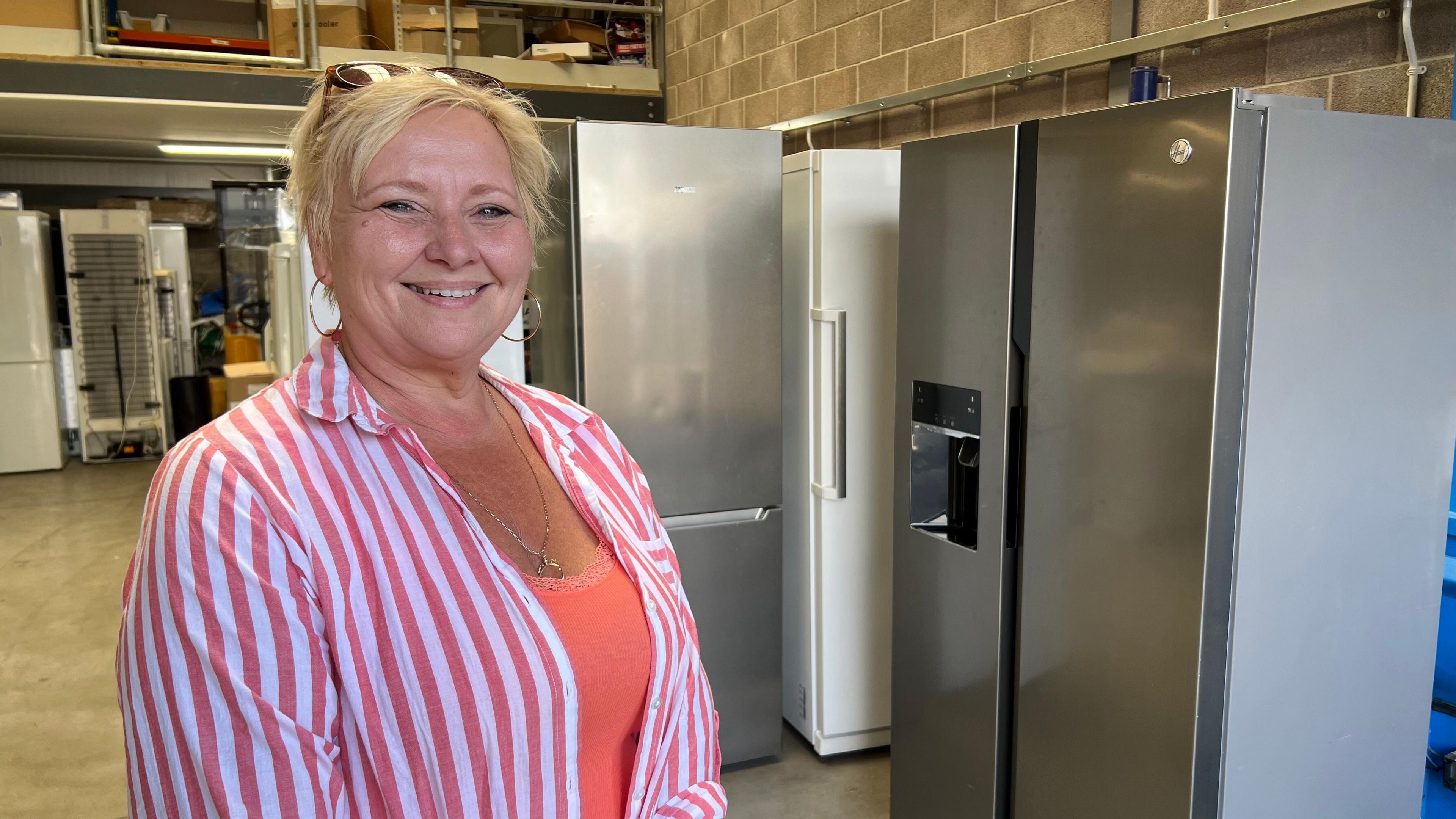 A woman stands in a pink and white striped shirt, in front of several fridges in a warehouse. She wears a coral coloured top under her shirt, and is smiling, with blonde hair.