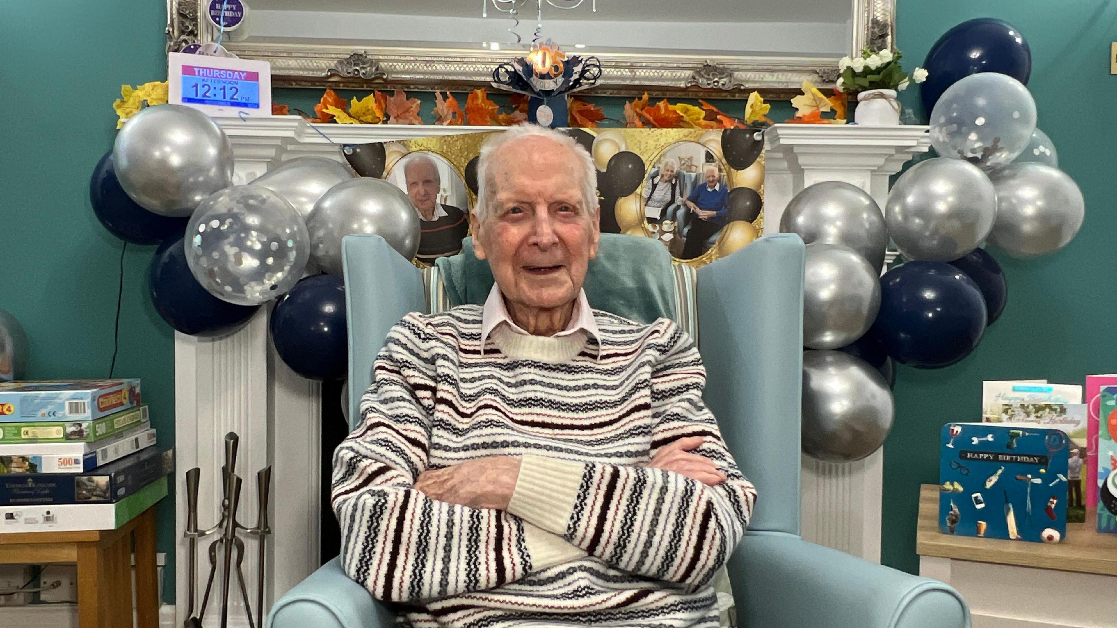 105-year-old Robert Warners. He is sat in a chair with his arms folded. Around him are blue and silver balloons and birthday cards. There is also a stack of board games his the left.