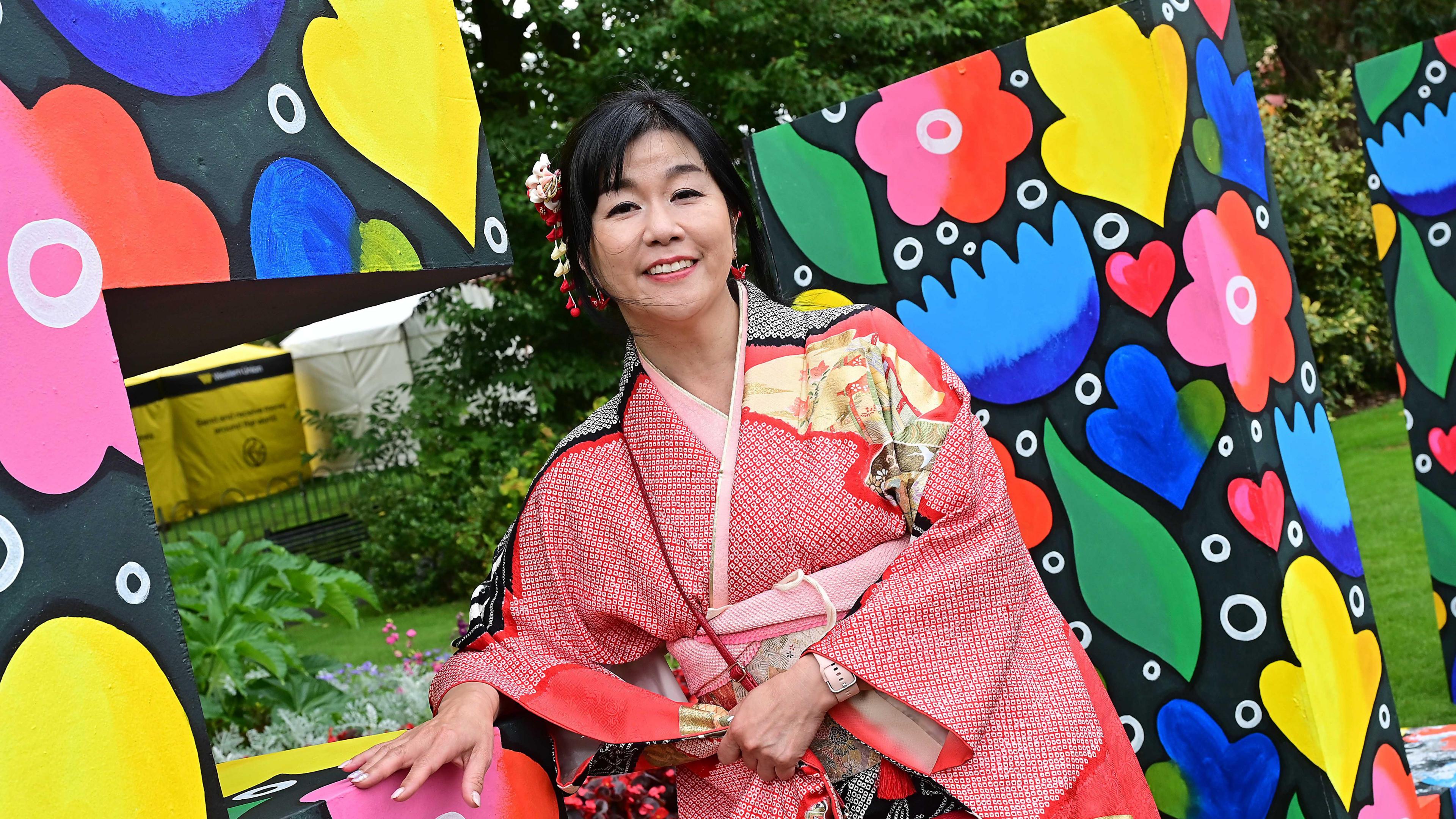 Women standing in red traditional outfit. Behind her is a background with different colours and green trees. 