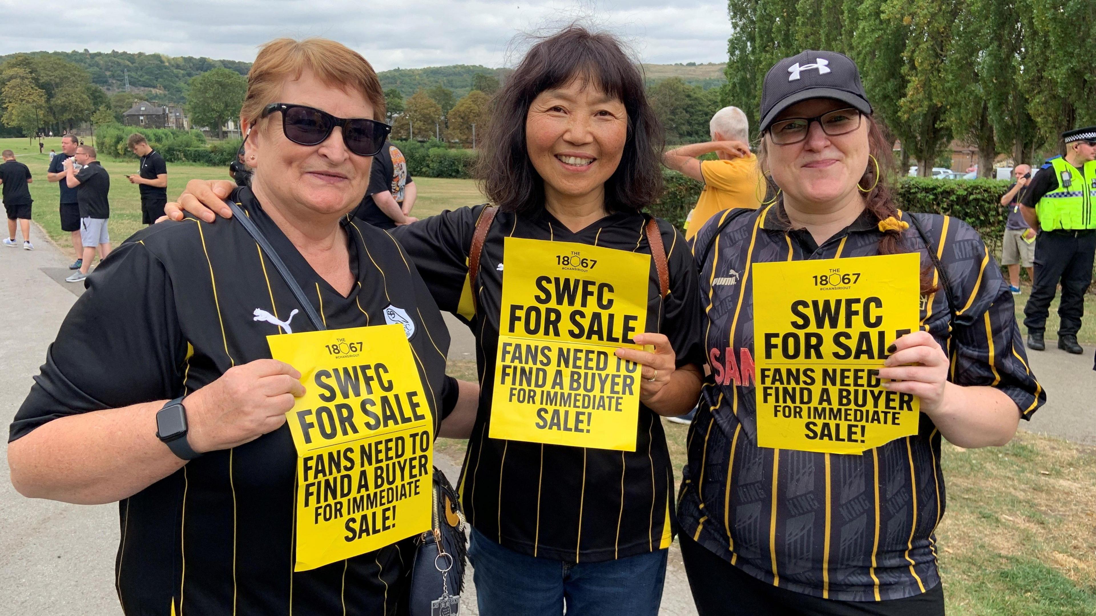 Three women are wearing black and yellow striped Sheffield Wednesday shirts. They are stood in a park holding yellow and black signs which say "SWFC for sale"