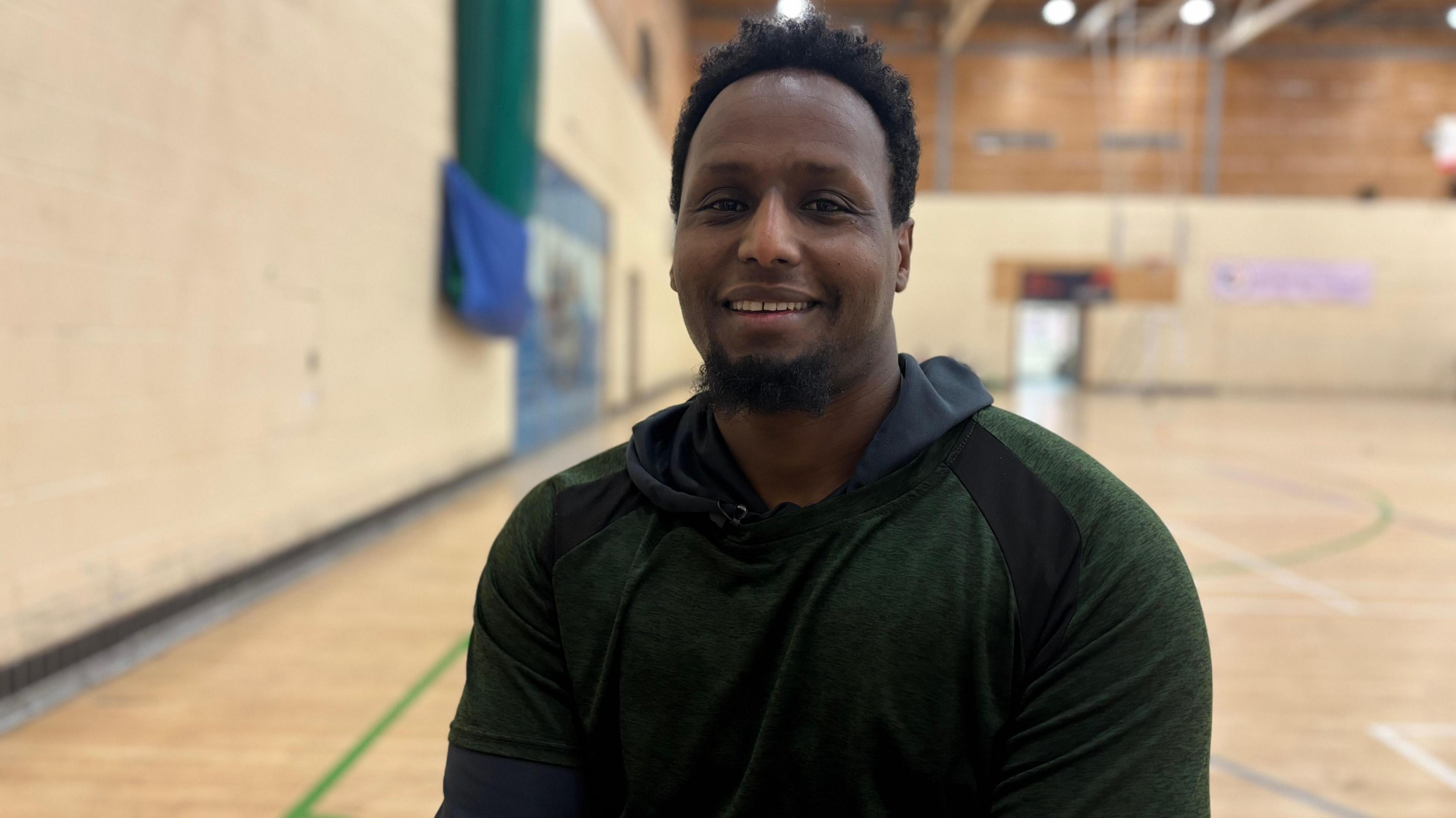 Abdi Jama sits in a sports hall. He has short dark hair and a short dark beard. He wears a green jumper