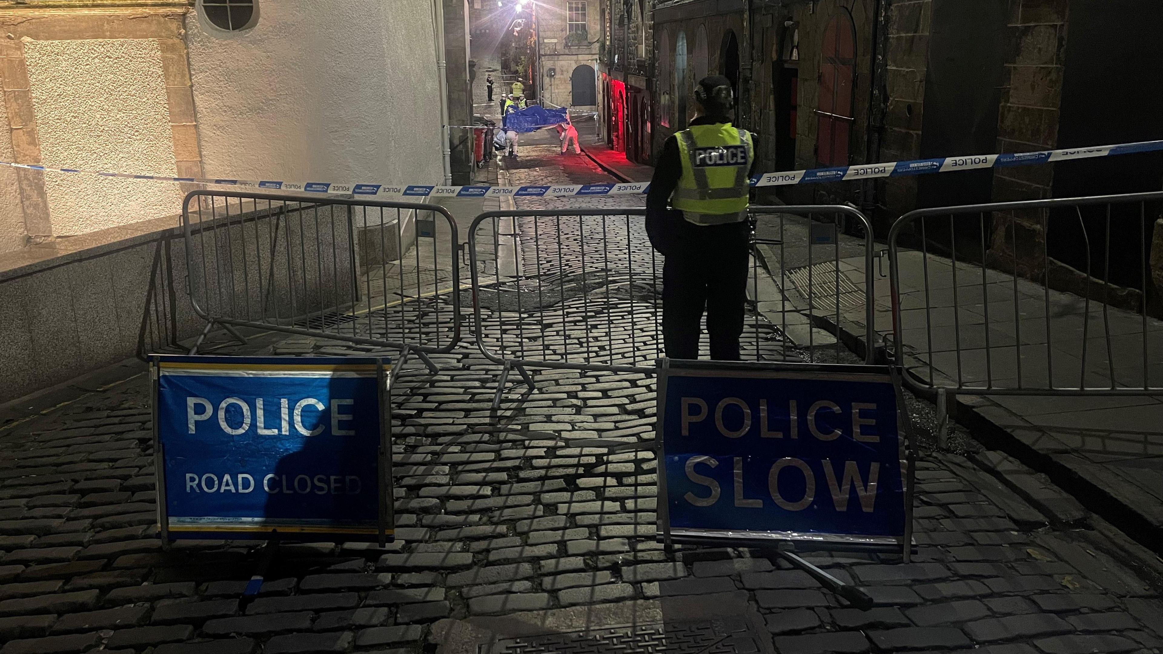 A police cordon on Niddry Street restricting access to the Cowgate. The street is cobbled. On one side is a pavement. On the other is a wall. Two blue signs are in the road. Both have white writing. One reads 'Police, Road closed'. The other says 'Police slow'. An officer wearing a flourescent vest over a dark uniform stands in front of a metal barrier and blue and white police tape. In the background, officers in white forensic overalls can be seen erecting a blue tent.