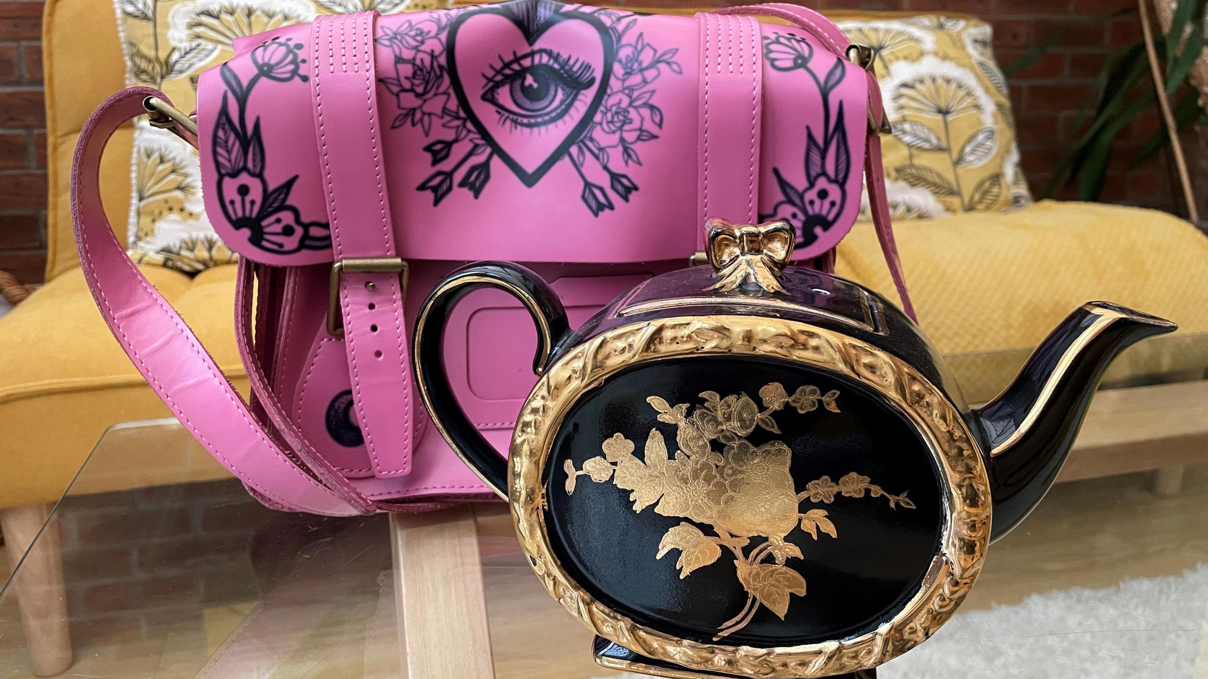 A pink Dr Martens bag with a flower pattern with an eye on it. It is next to a black and gold teapot, in front of a yellow sofa with two cushions on it.