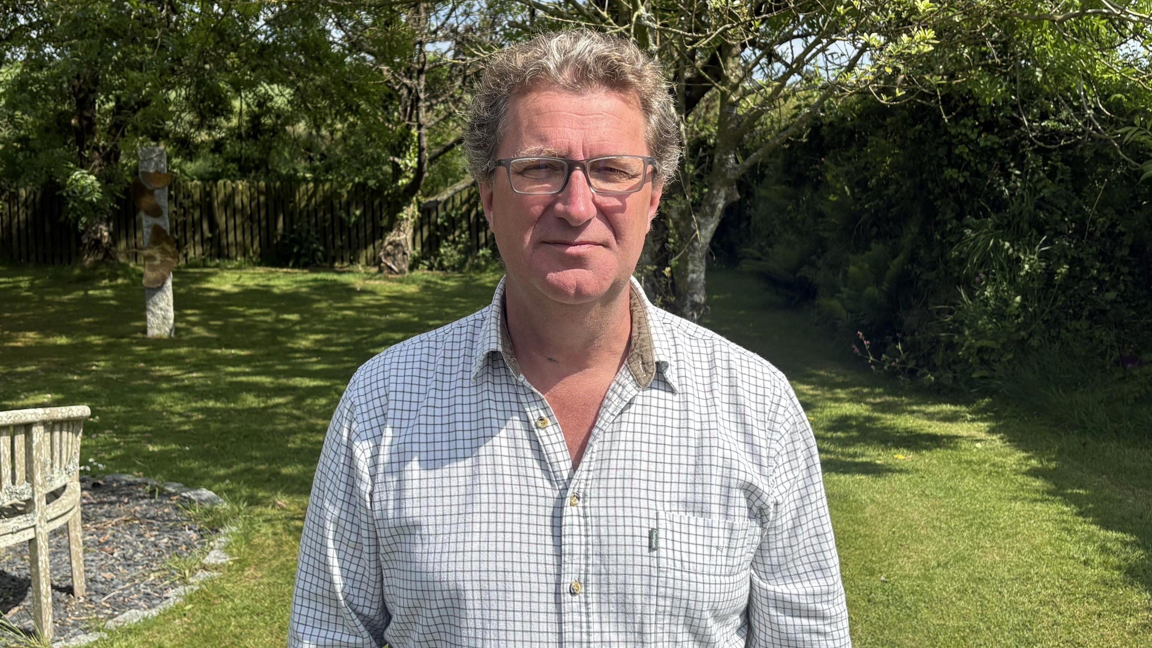 A man wearing a white checked shirt. He is wearing glasses and has curly greying hair. He is standing on a lawn with trees in the background.