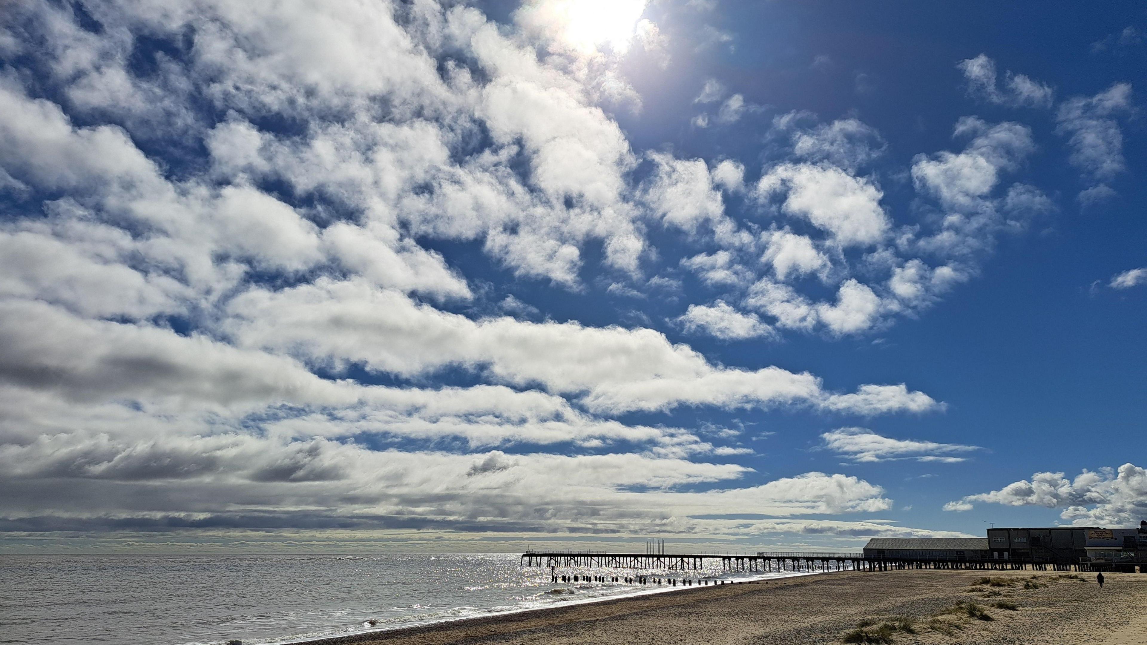 Sand and shingle beach looking out to sea with a pontoon in the background under a blue and cloudy sky with sun peering through