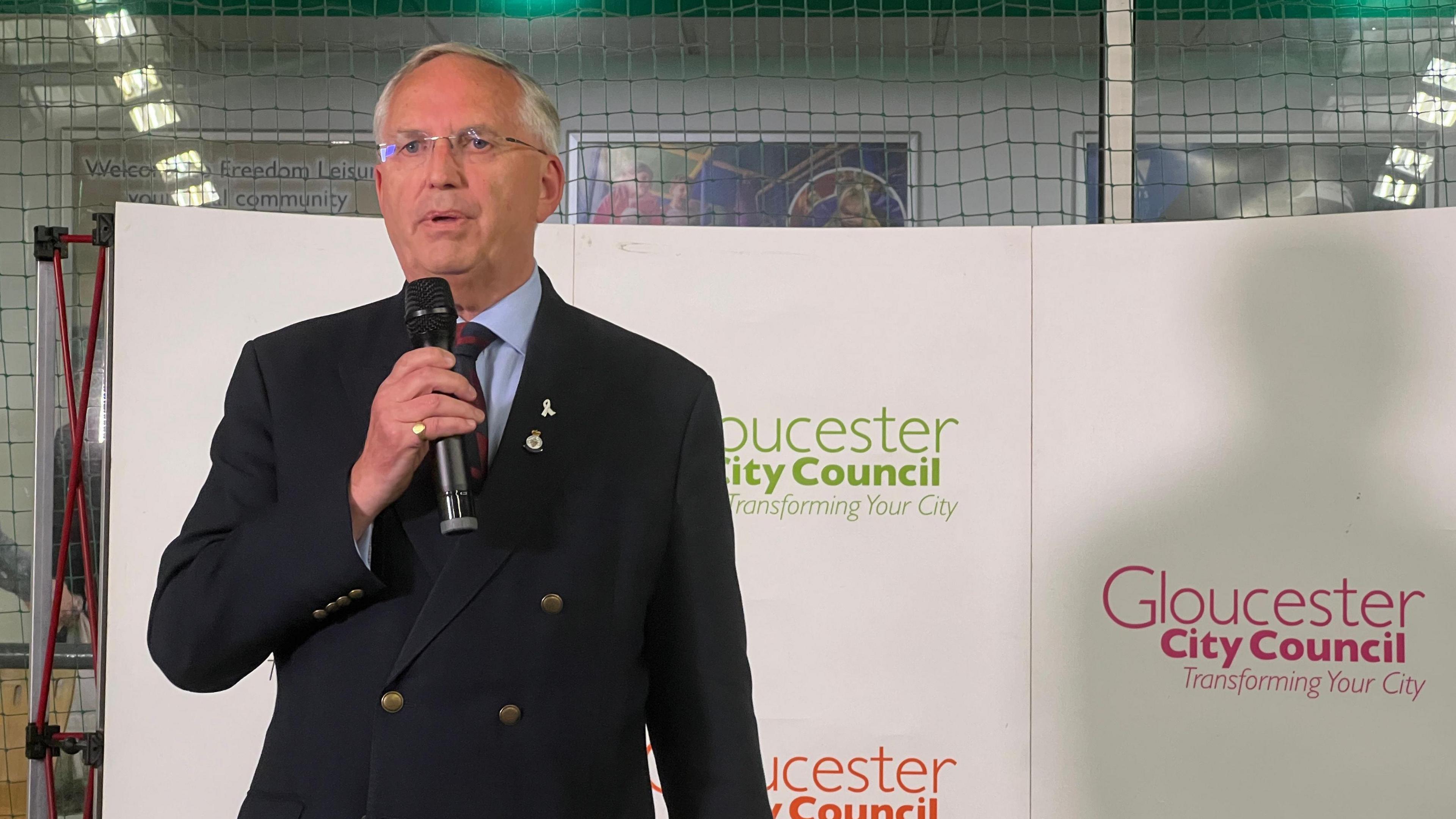 Chris Nelson speaking into a microphone against a Gloucester City Council backdrop