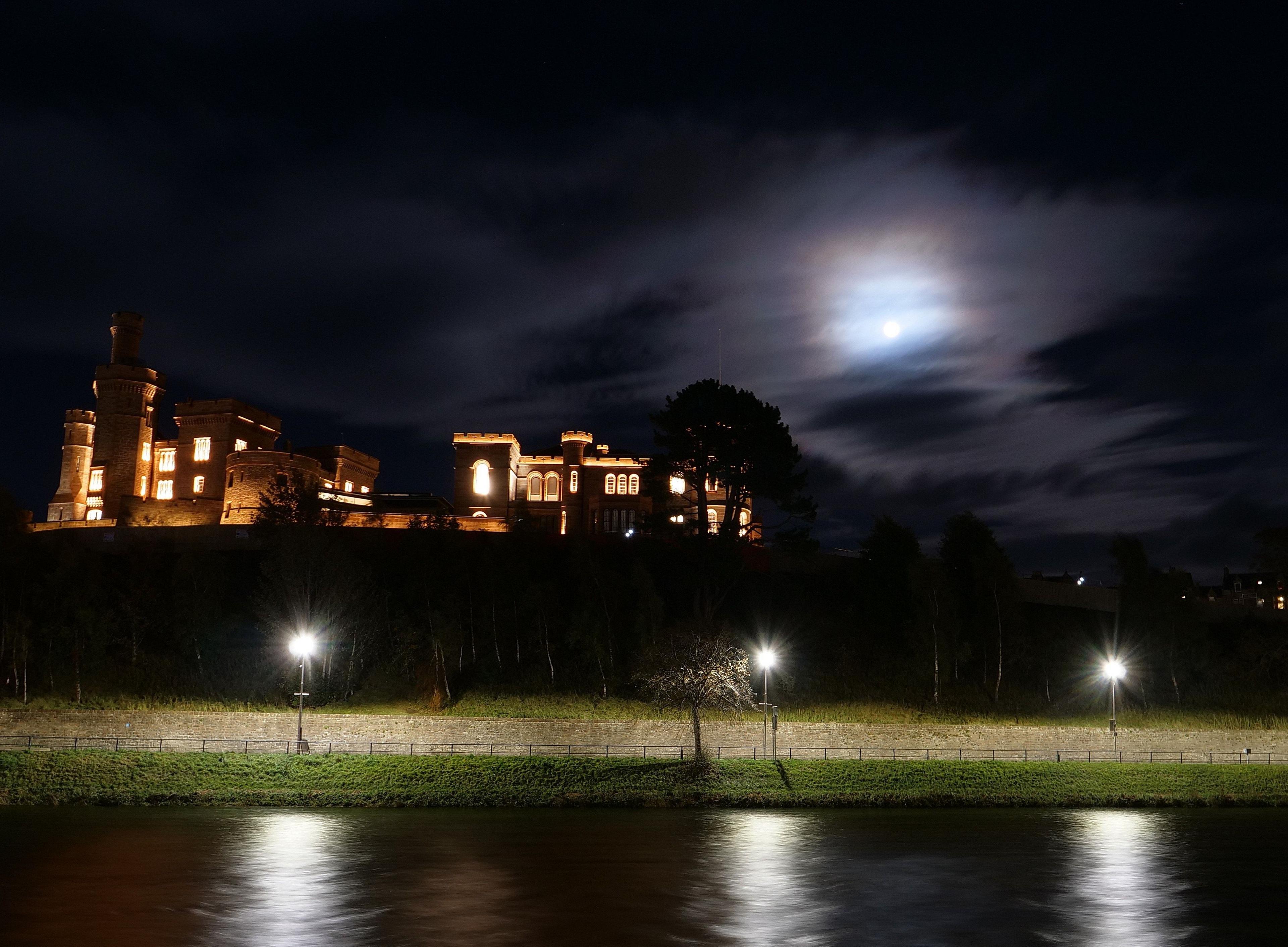Brightly lit castle on a hill, below a dark cloudy sky with a bright, full moon peeking through the clouds.