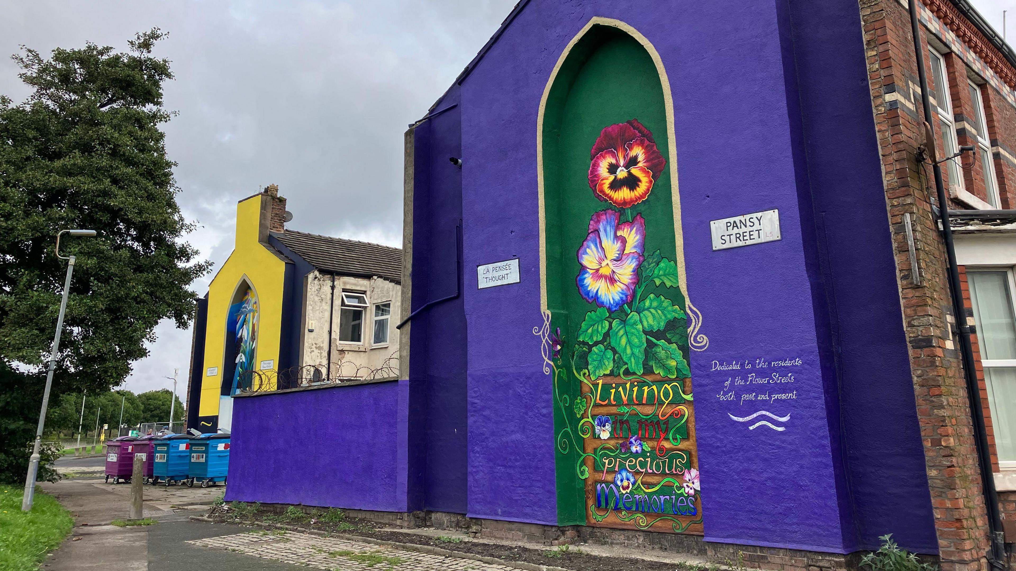 A gable-end of Pansy Street is painted purple, with a green section in the centre featuring a giant pansy and the words 'Living in my precious memories' and 'Dedicated to the residents of the Flower Streets both past and present'.