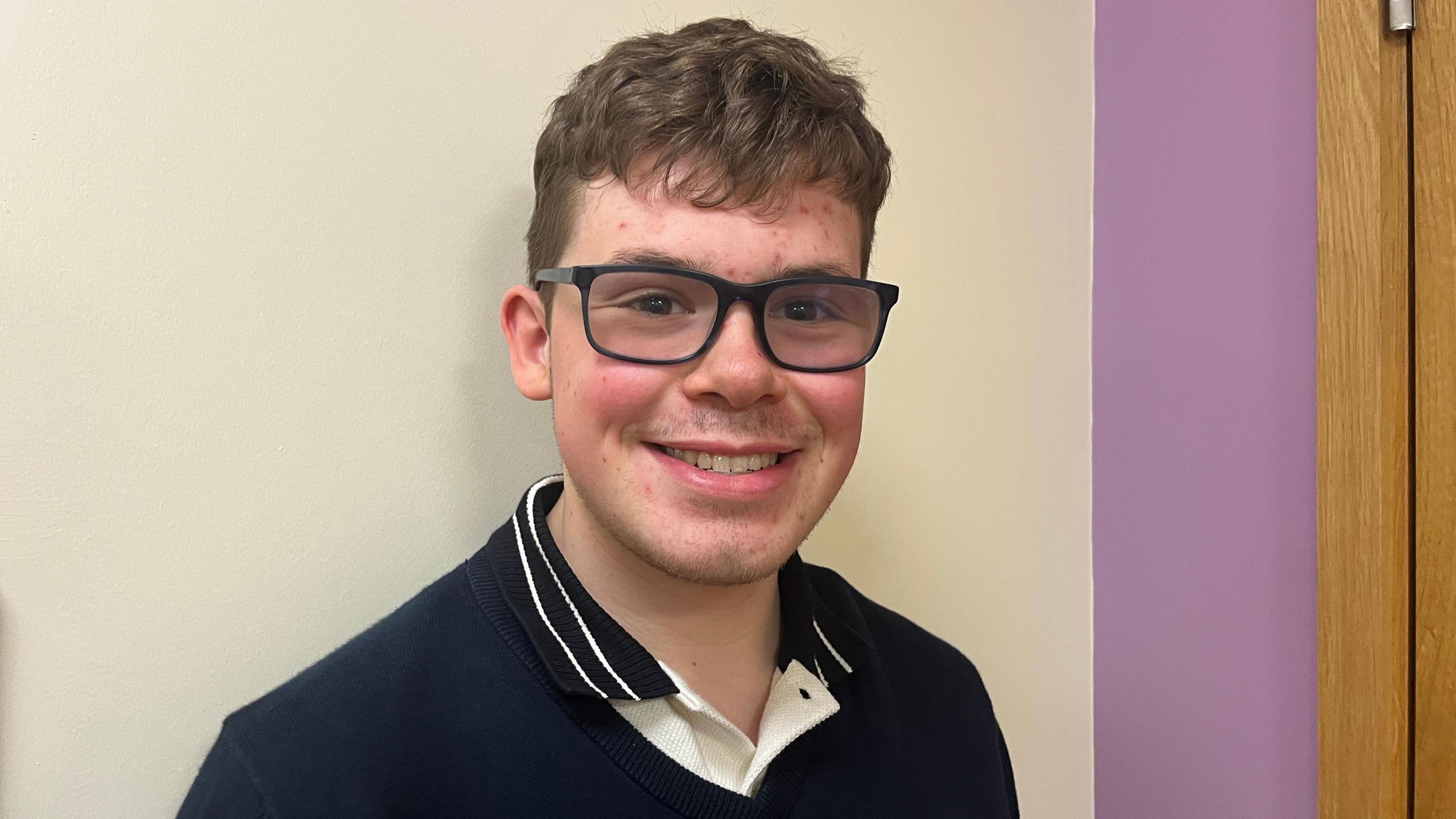 Man looking into the camera smiling. He has short, brown, curly hair and is wearing thick black glasses which are tinted slightly. He is wearing a blue polo shirt. In the background, the wall is white with a purple strip to the right. 