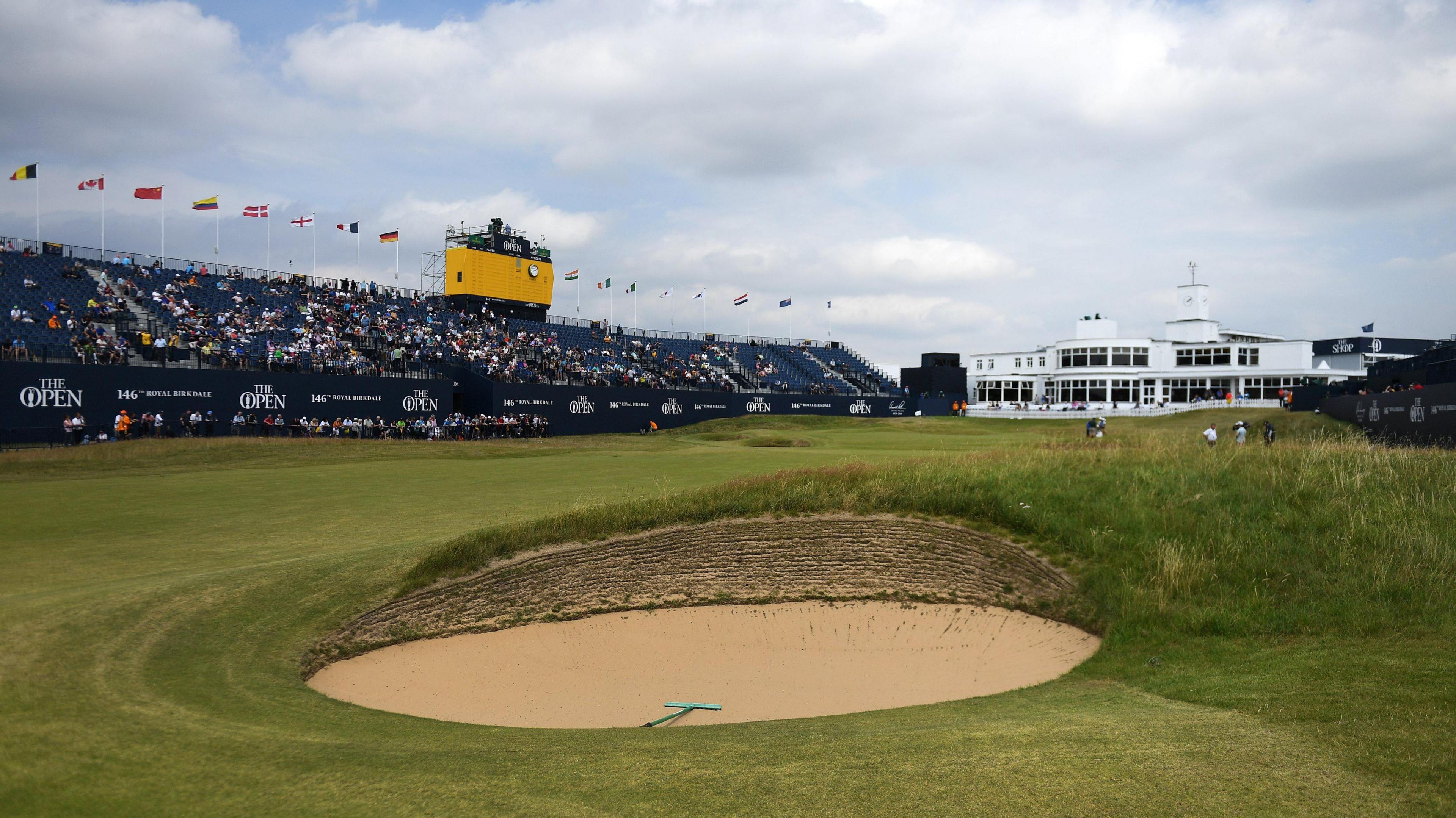 a golf course, with a sandy bunker in the foreground. On the left there is a large stand, approximately half full of fans. Above that, there is a small TV gantry for cameras. To the right of the stand, a large white building is pictured. On the course, there are several golf players and caddies in the distance.