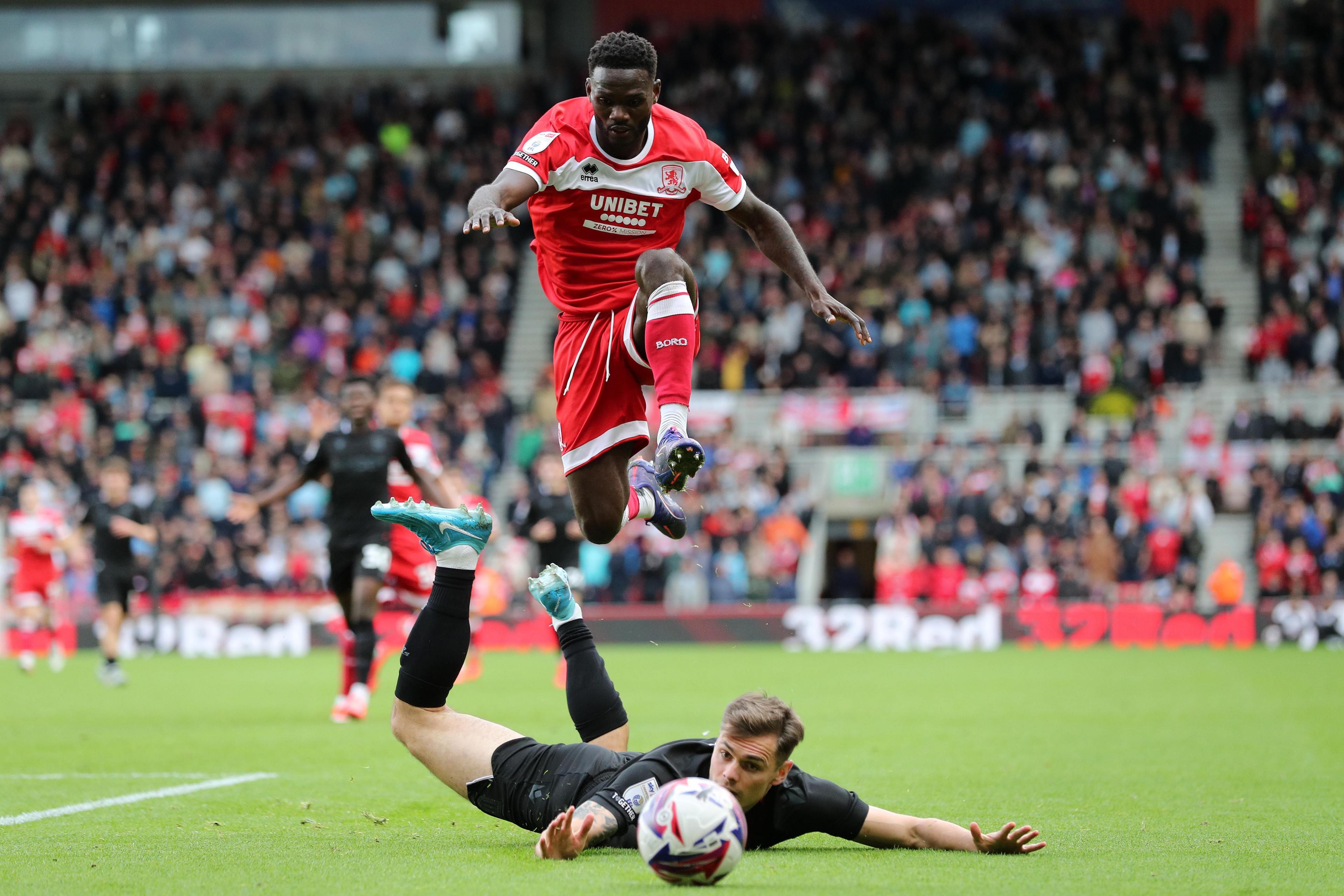 A Middlesbrough player in all red chases a ball as he jumps over another player all in black lying on the ground.