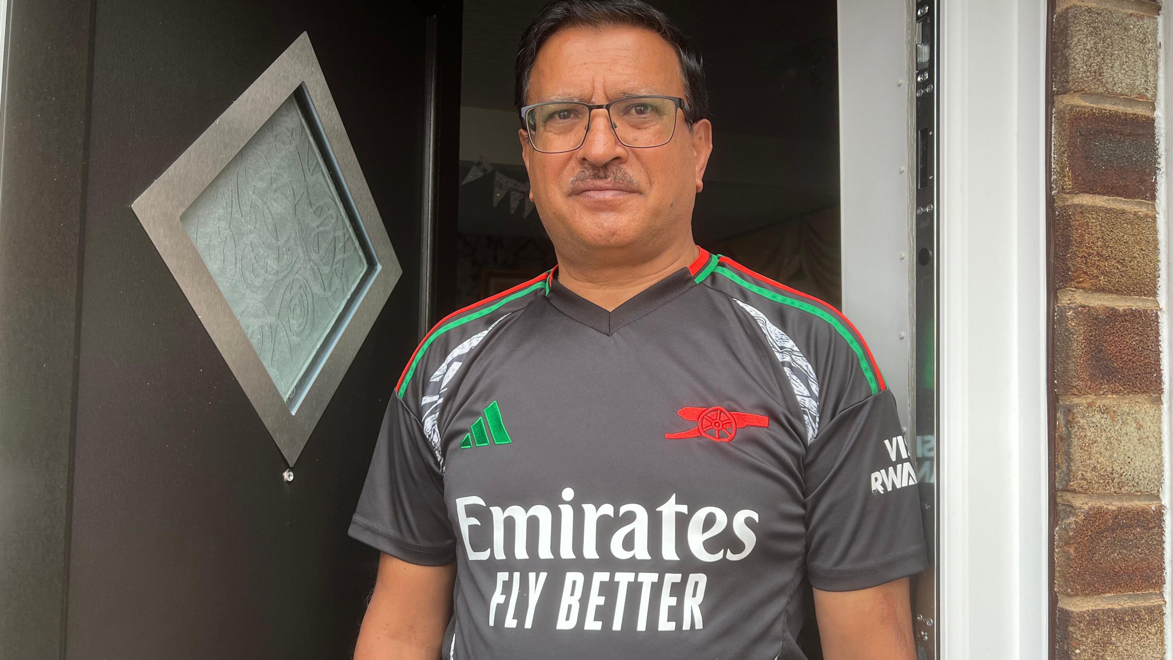 A middle aged man of South Asian heritage stands in a front doorway. He wears a grey football shirt with green and red branding.