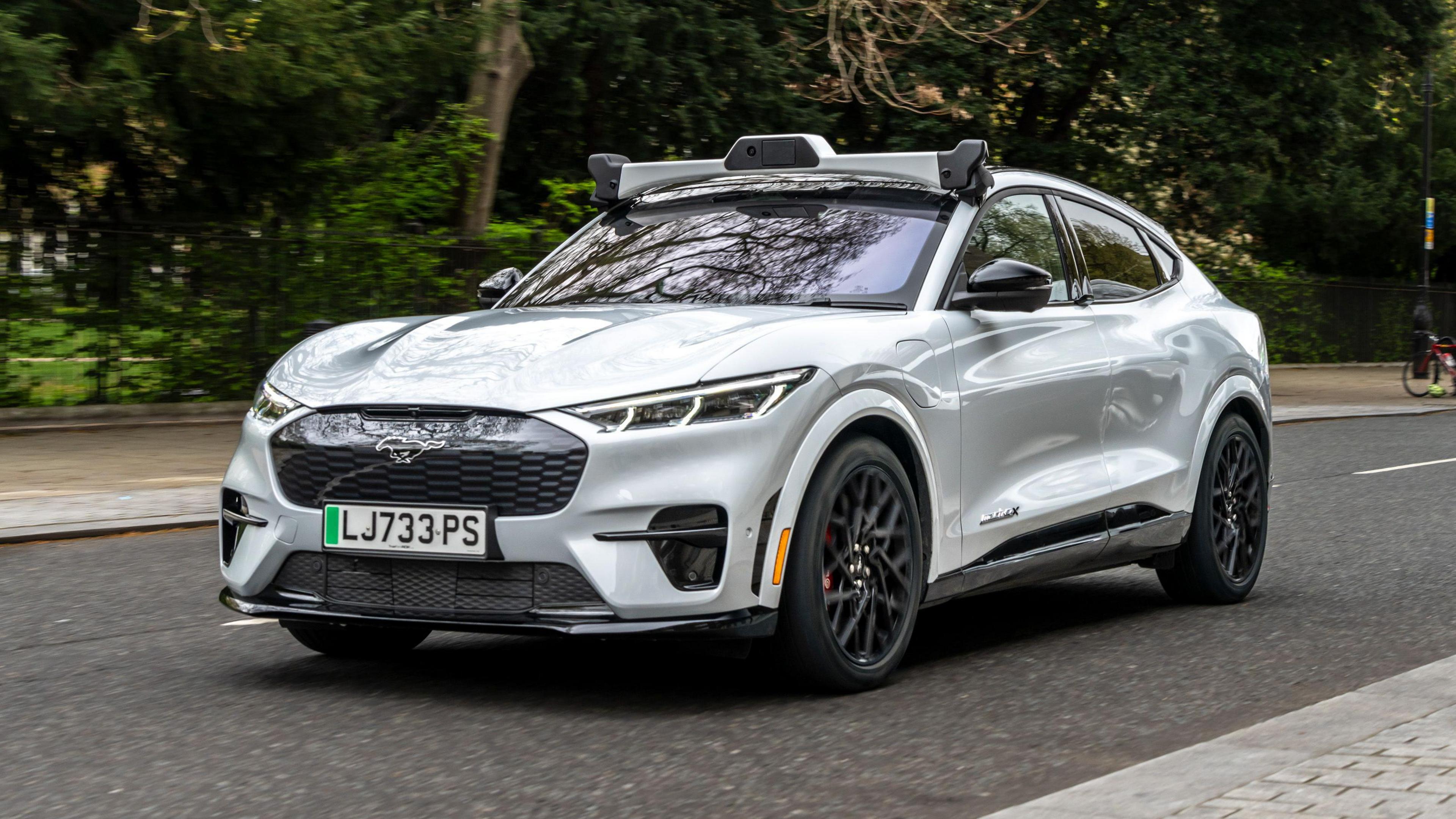 A silver electric vehicle is shown driving down a street in London. It is driving past a park, with green trees in the background.
