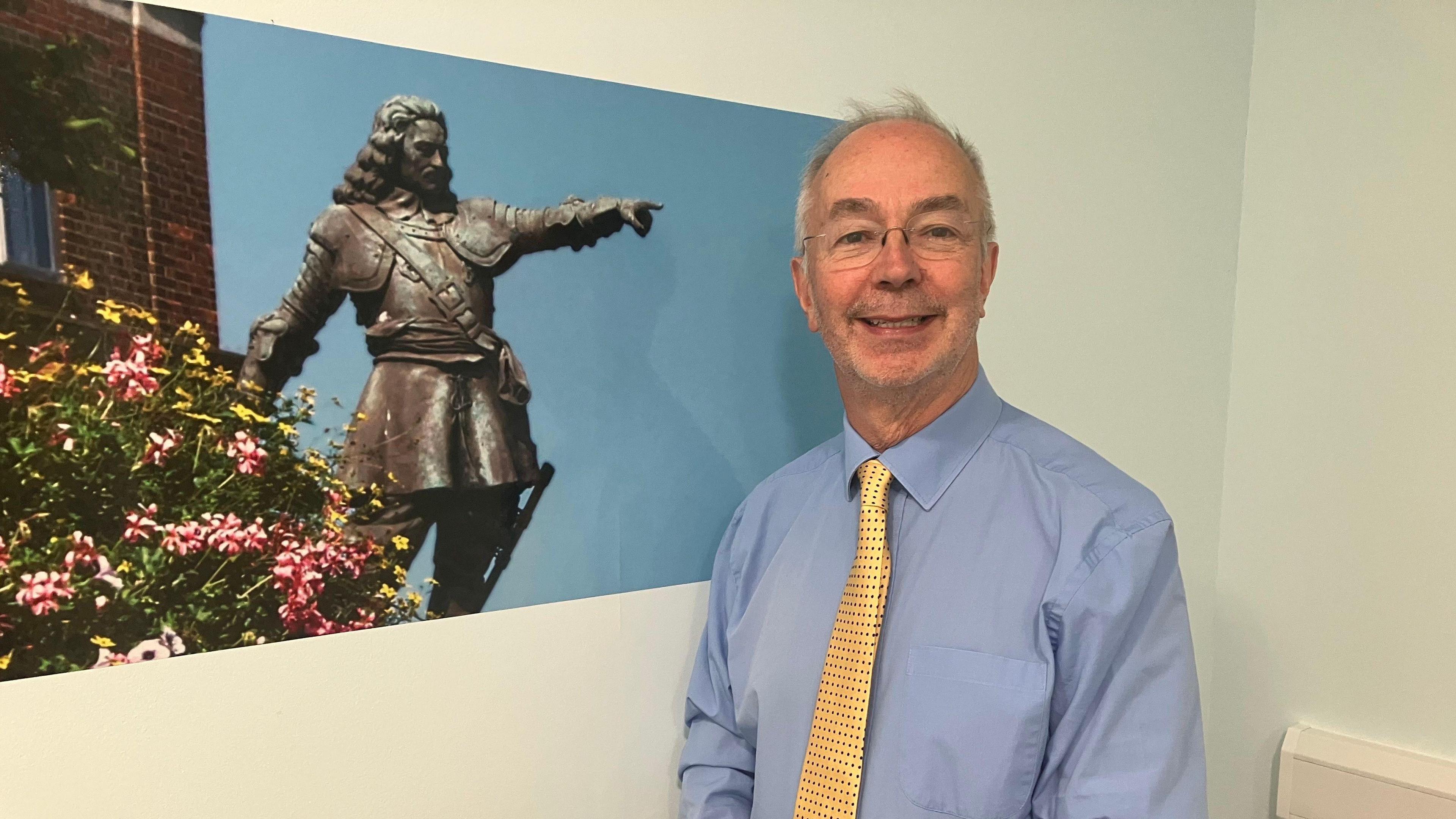 A picture of Buckinghamshire Council leader Martin Tett. He is stood next to a picture of historical figure John Hampden. He is wearing a yellow tie with black dots on it and is smiling.