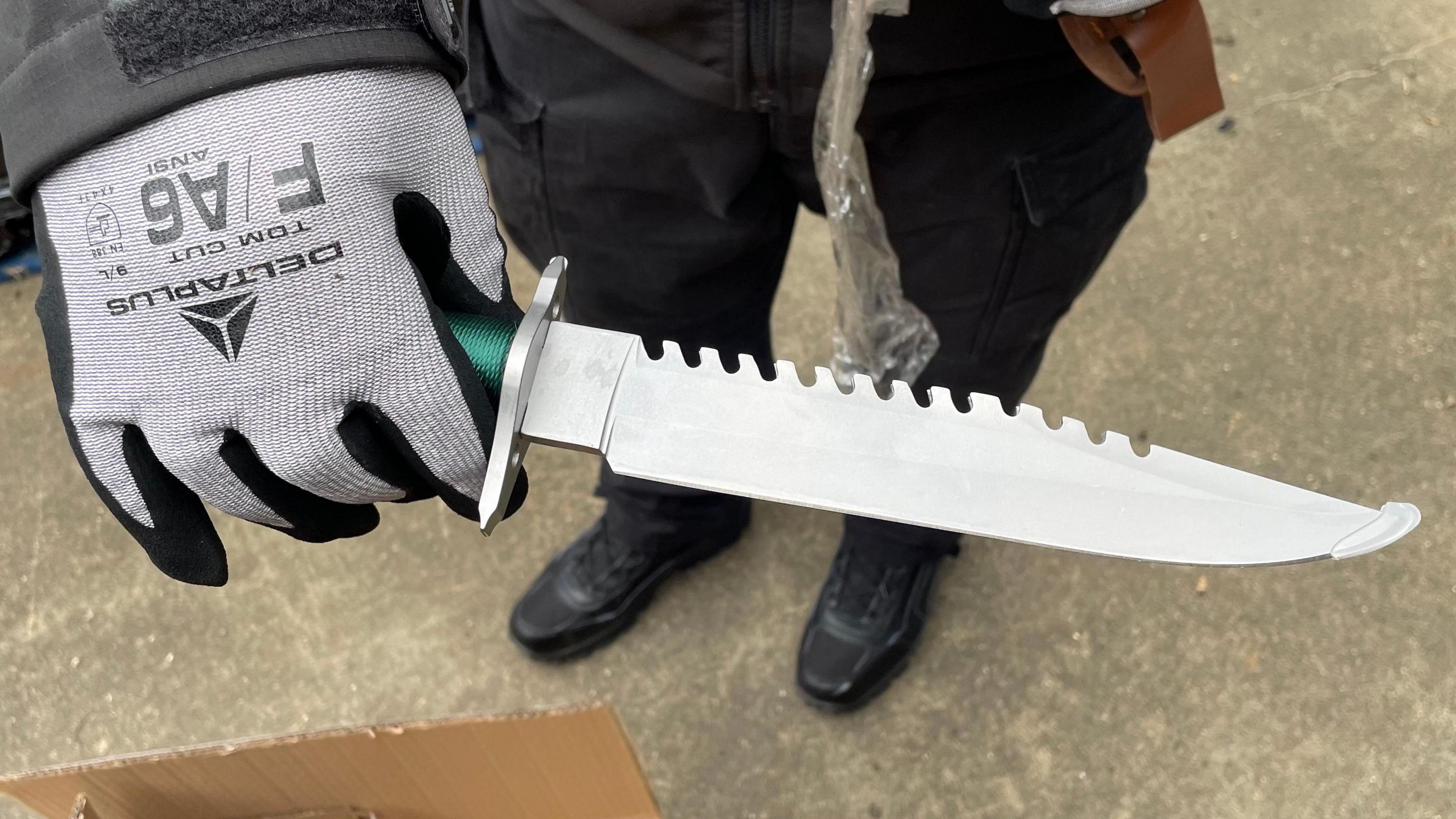 A gloved hand holding a knife. The blade is more than 8in (20cm) long and is sharpened on one side, with serrations on the other. A small part of a handle wound with green string is visible. The hilt and the blade are both bright, new metal. The tip of the blade is covered with a small piece of safety plastic. In the background are the legs and feet of the police officer holding the knife, standing on a concrete surface.