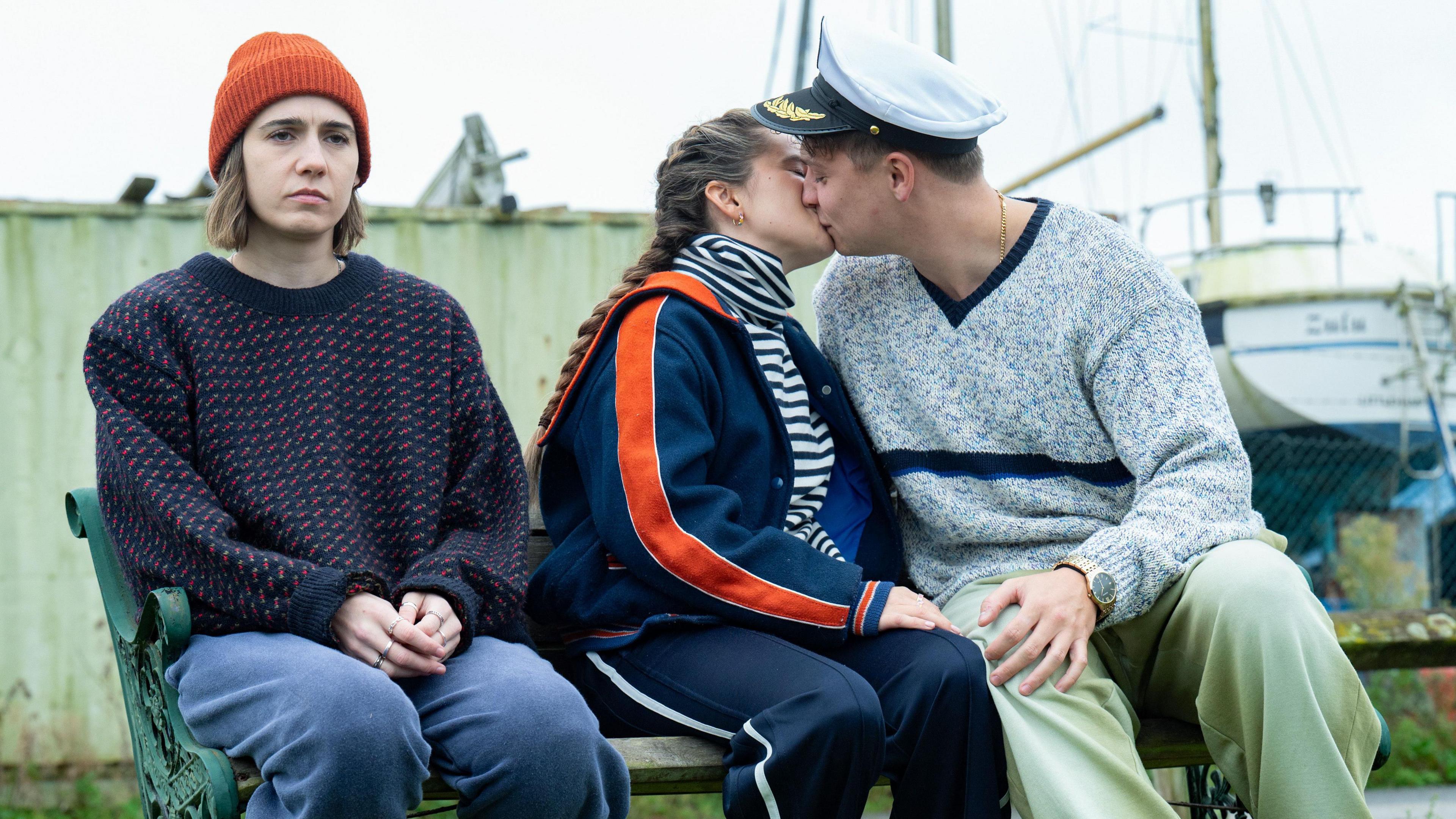 From left to right: Sitting on a bench Josie (Sadler) is wearing blue trousers and a blue jumper with an orange beanie. She is sitting next to Billie (Davidson) wearing a blue tracksuit with a striped top and is kissing Nicky (Sam Buchanan) who is wearing green trousers a grey jumper and a sea captain's hat.