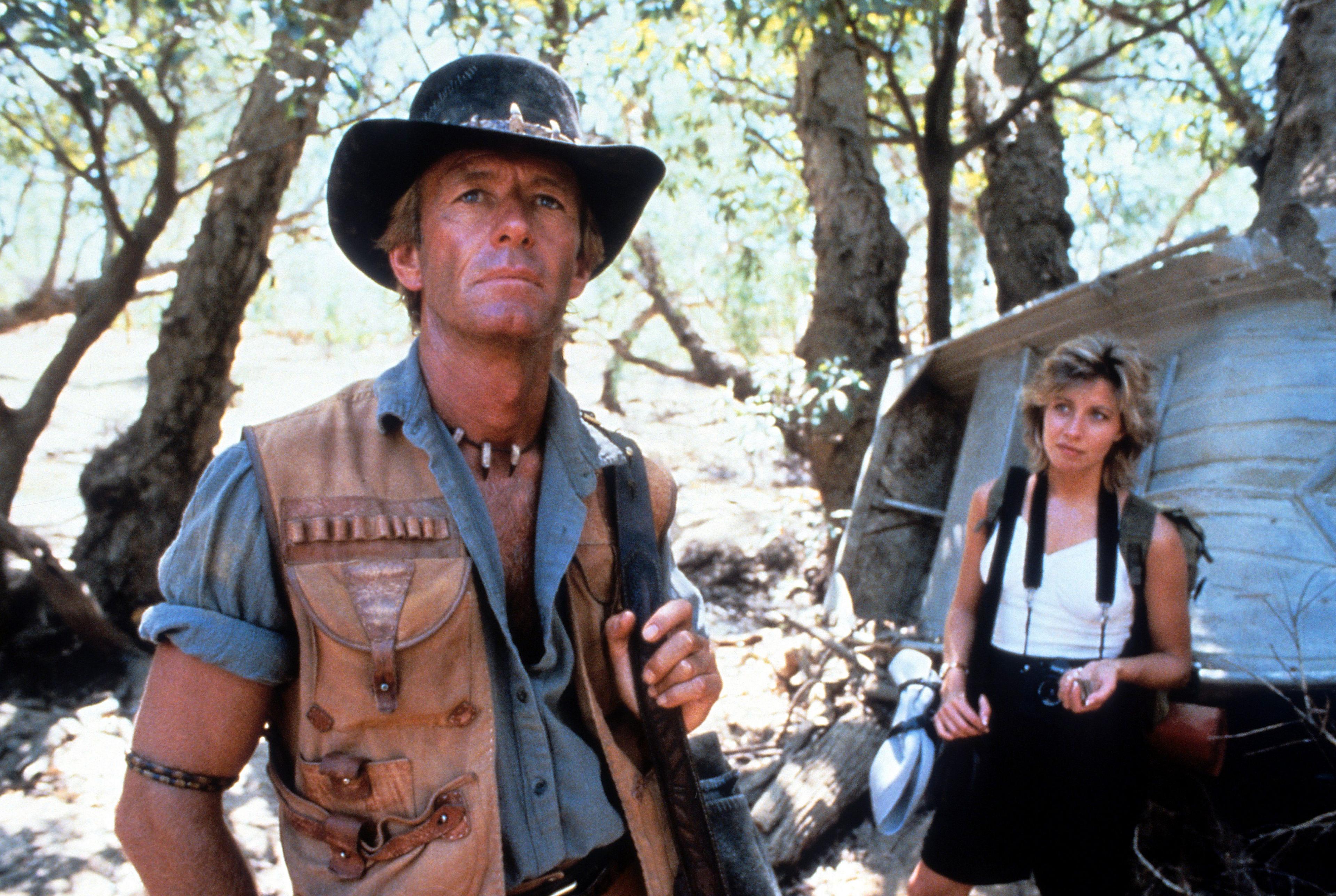 Paul Hogan in the foreground wearing a hat, denim shirt and tan-coloured waistcoat. Linda Kozlowski is in the background, wearing a white vest and black shorts.