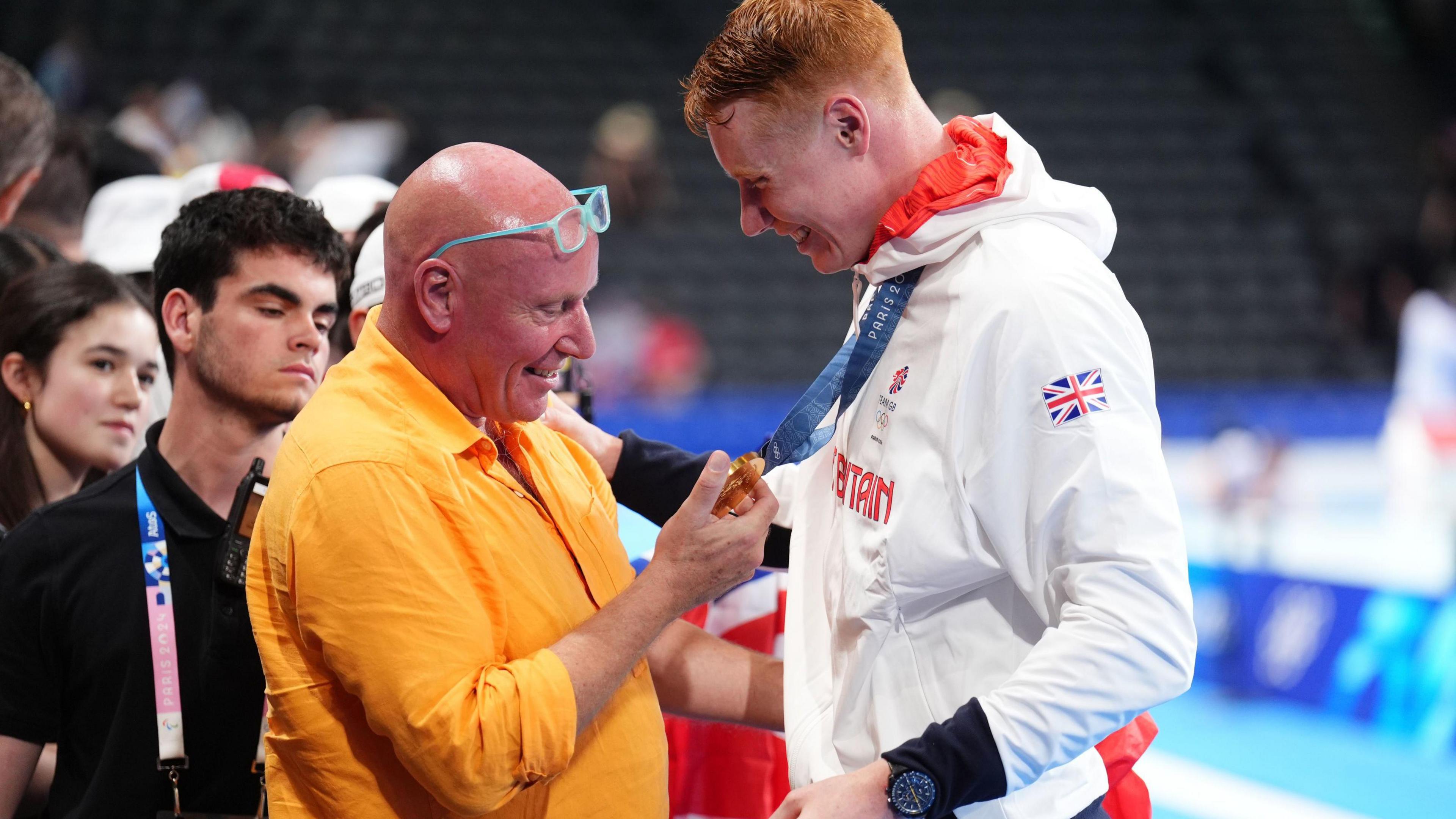 Tom Dean, in his Team GB tracksuit, is on the right, with his dad, Jonathan, who is bald and wearing an orange shirt and has blue-rimmed glasses perched on his head, is smiling as he looks at the medal