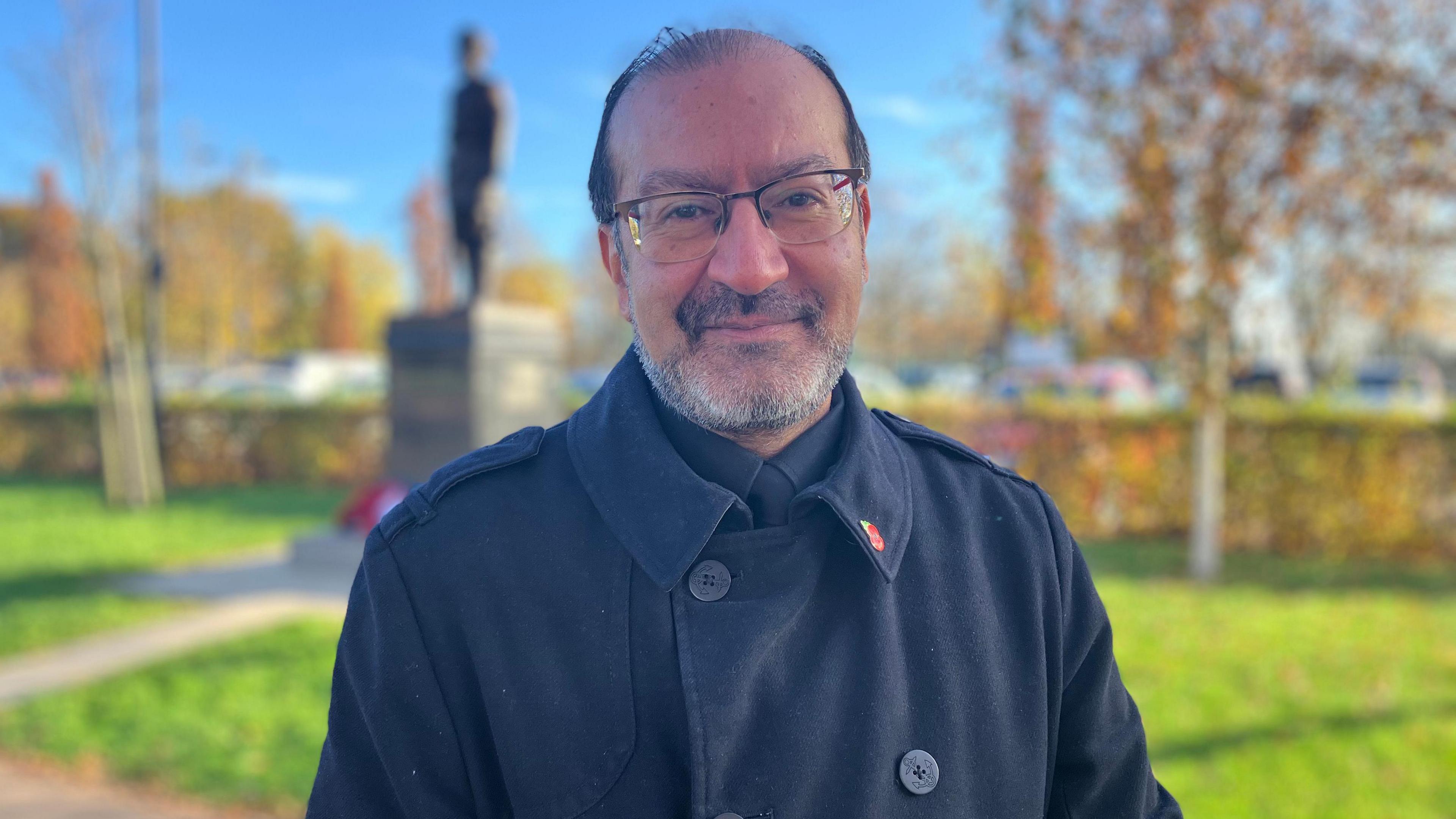 Gurinder Singh wearing a black coat with a poppy pin badge and glasses. The statue of the Sikh solider can be seen behind him out of focus.