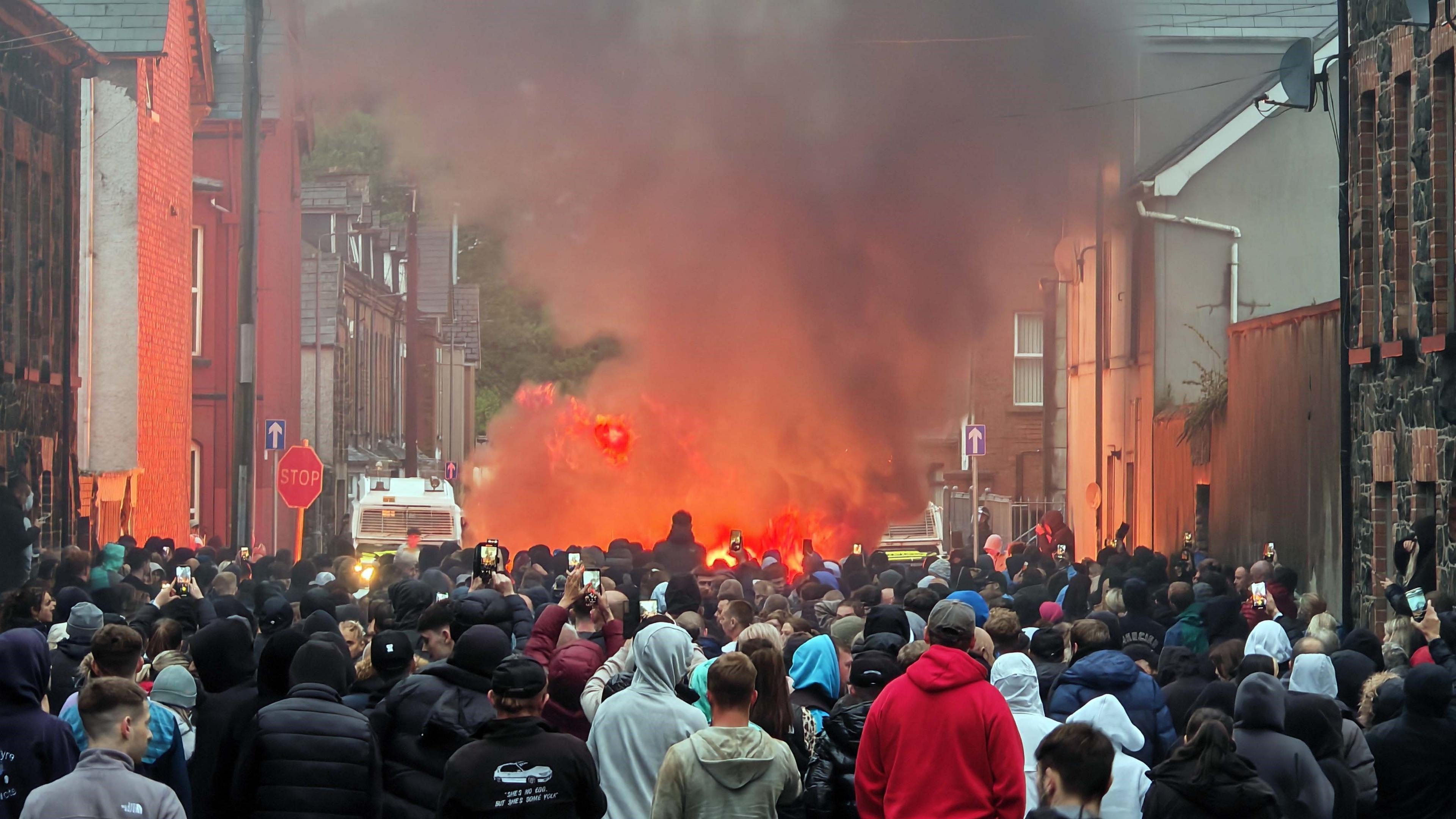 People on the streets during Ballymena protest. A large crowd. There are buildings along the side of the street and a police van in the distance. There is a white police van. Smoke is rising into the air.