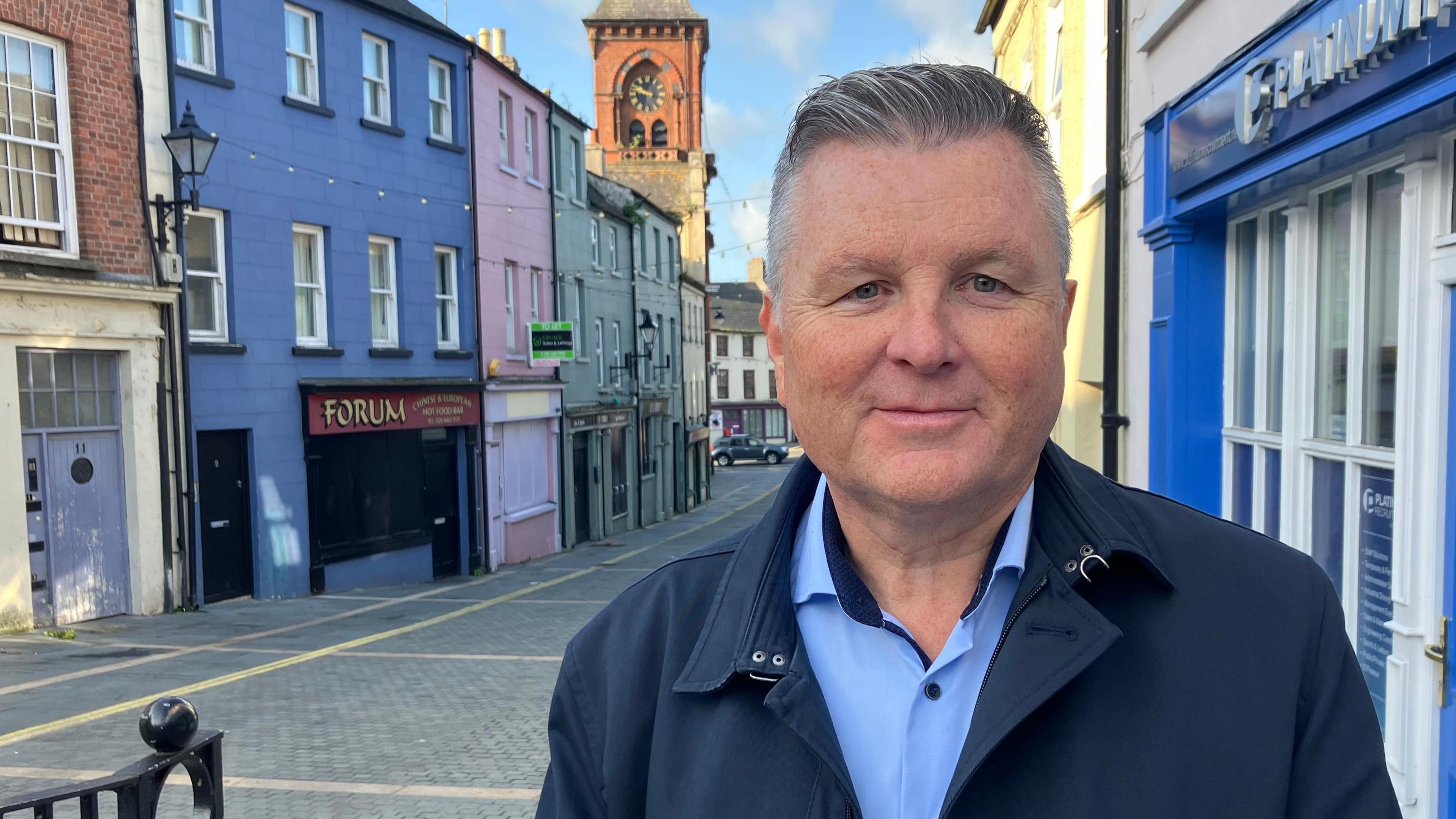 A man wearing a navy jacket with a light blue shirt underneath. He has grey hair is pushed back off his forehead. He is standing on a street with lots of colourful  buildings behind him.  