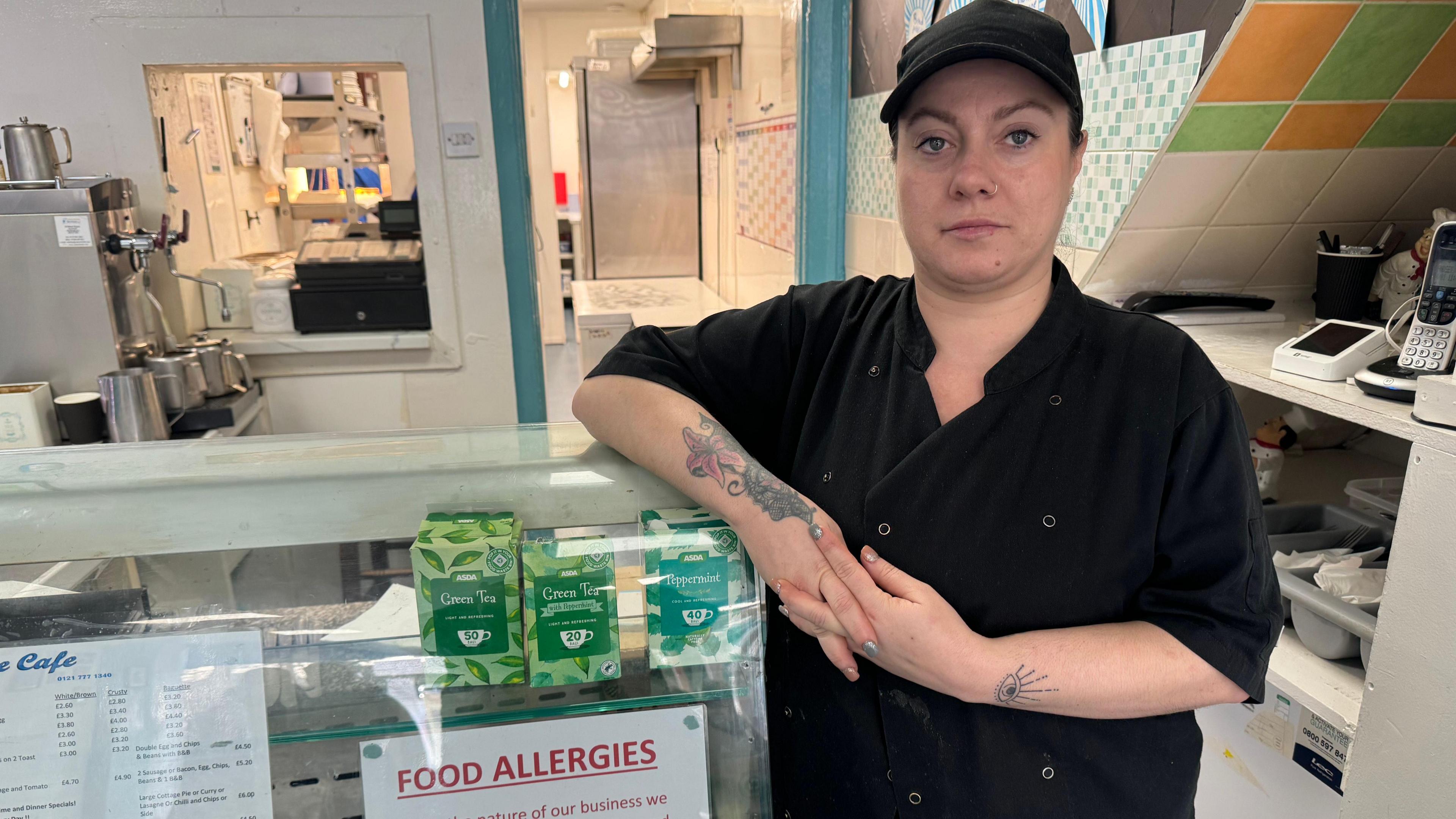 A woman wearing a black cap and shirt stands on the side of a counter staring into the camera. On the counter is a glass shield with a menu for the cafe on and a note about food allergies, next to green boxes of tea.