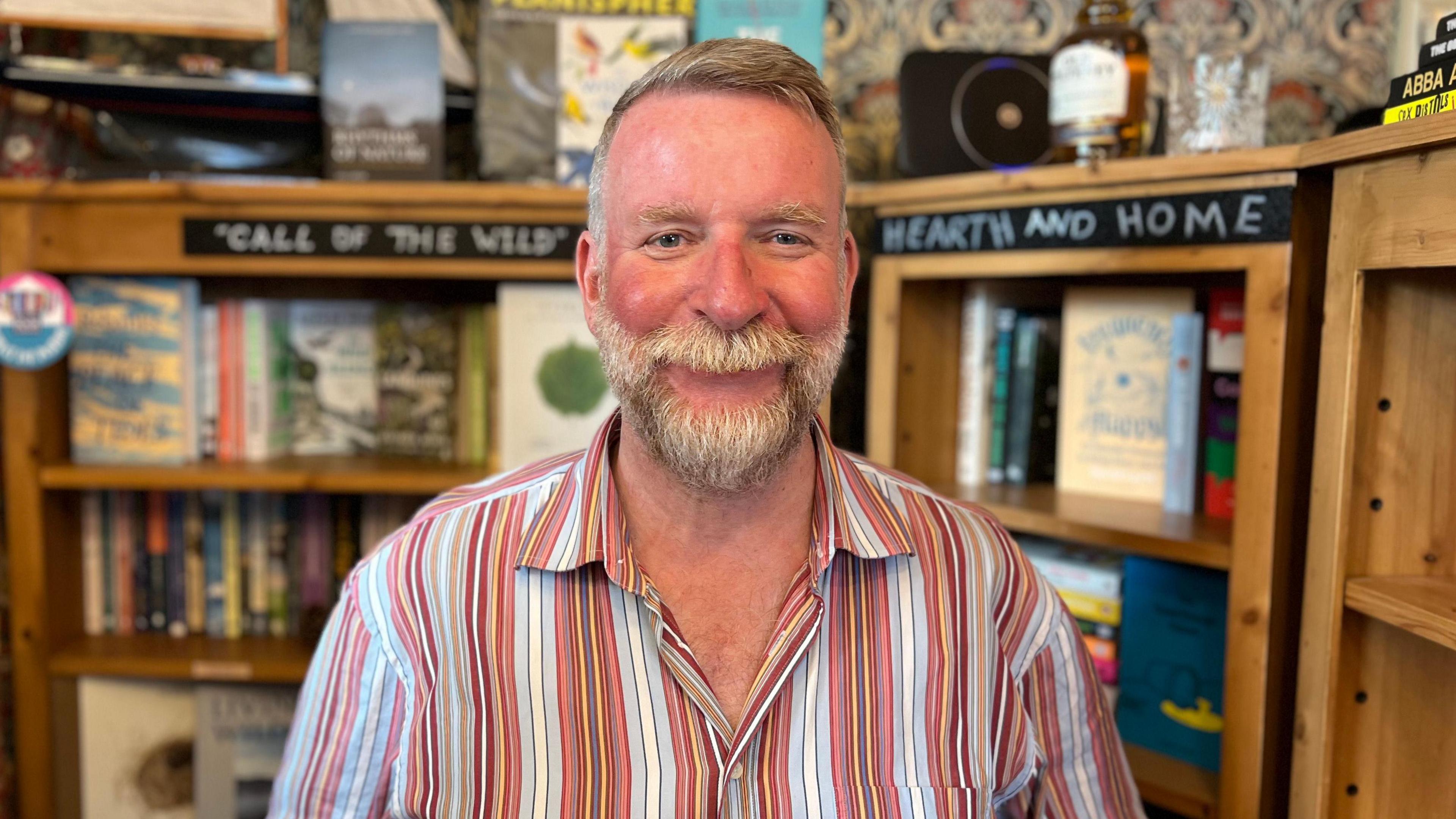 Stuart Parsons is smiling at the camera in his bookshop. He has short grey hair in a quiff and a medium length beard. He is wearing a colourful striped shirt featuring light blue, red, white yellow.